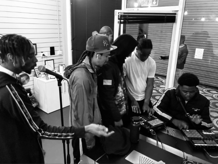 Black and white image of five people gathered around music equipment in a studio with horizontal blinds and white walls.