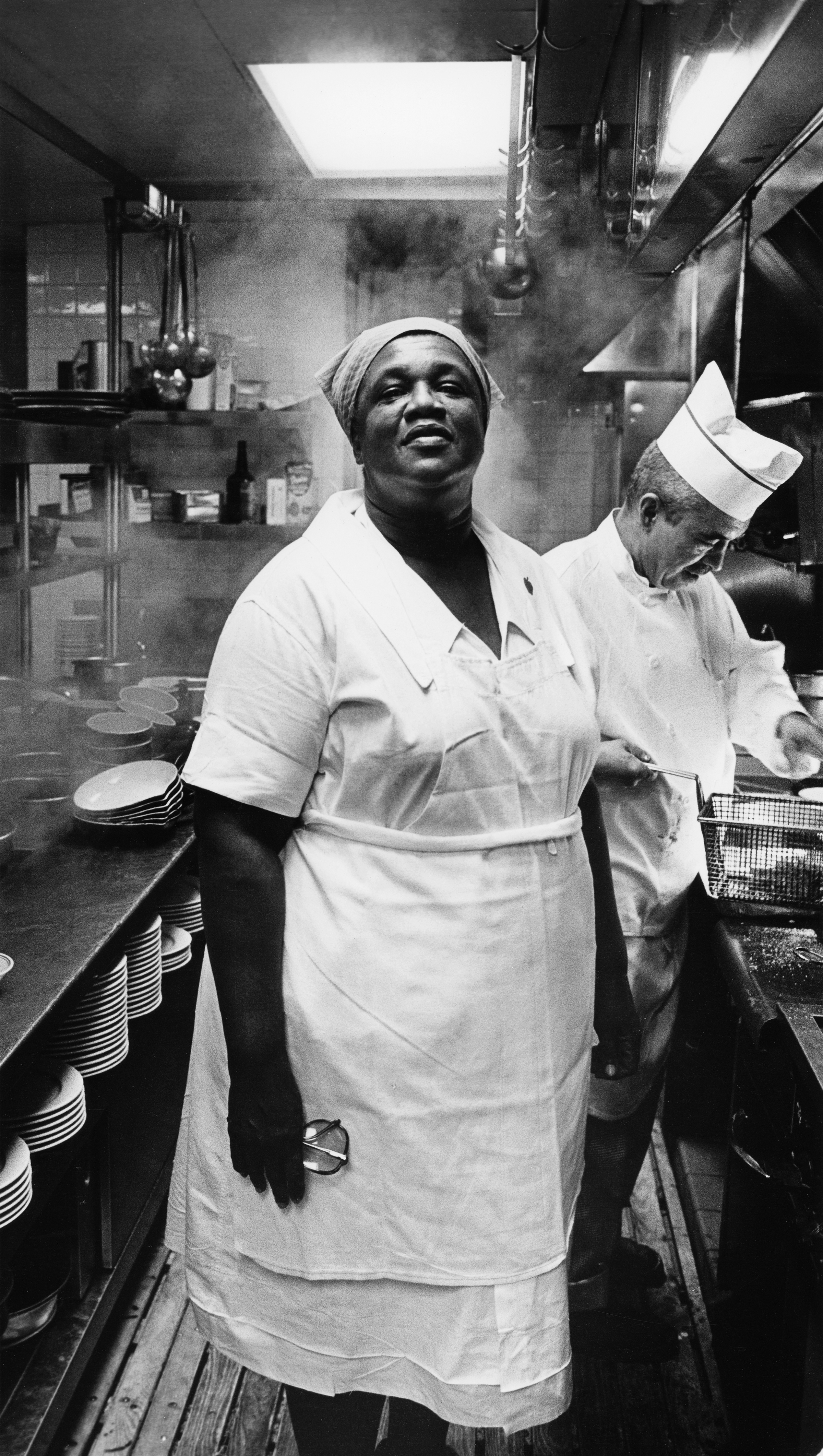 Black and white image of smiling woman in white chef's coat and headwrap standing in busy commercial kitchen with steam and equipment.
