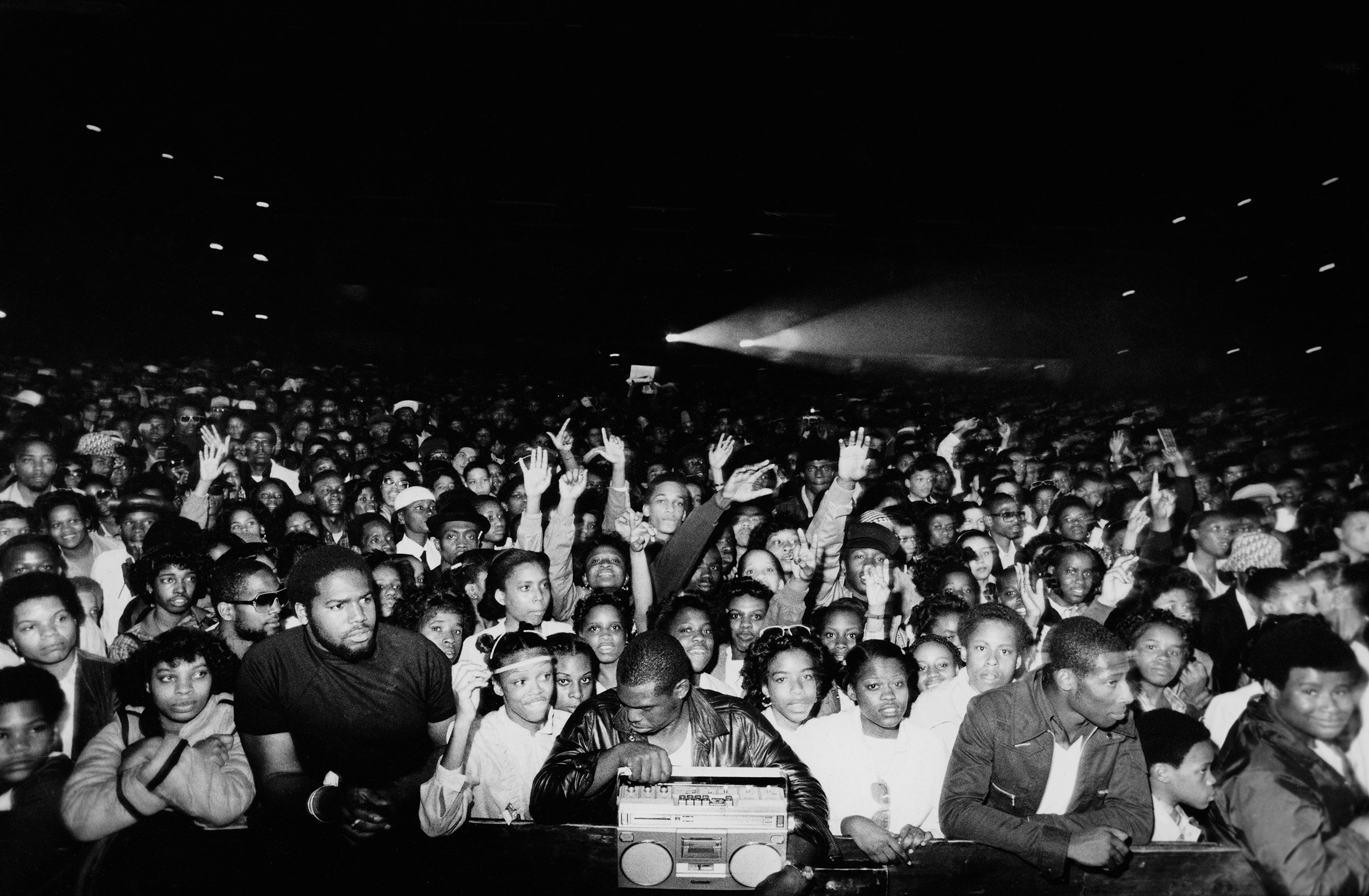 Black and white image of densely packed crowd with raised hands, many wearing caps and casual clothing, with boombox visible in foreground.