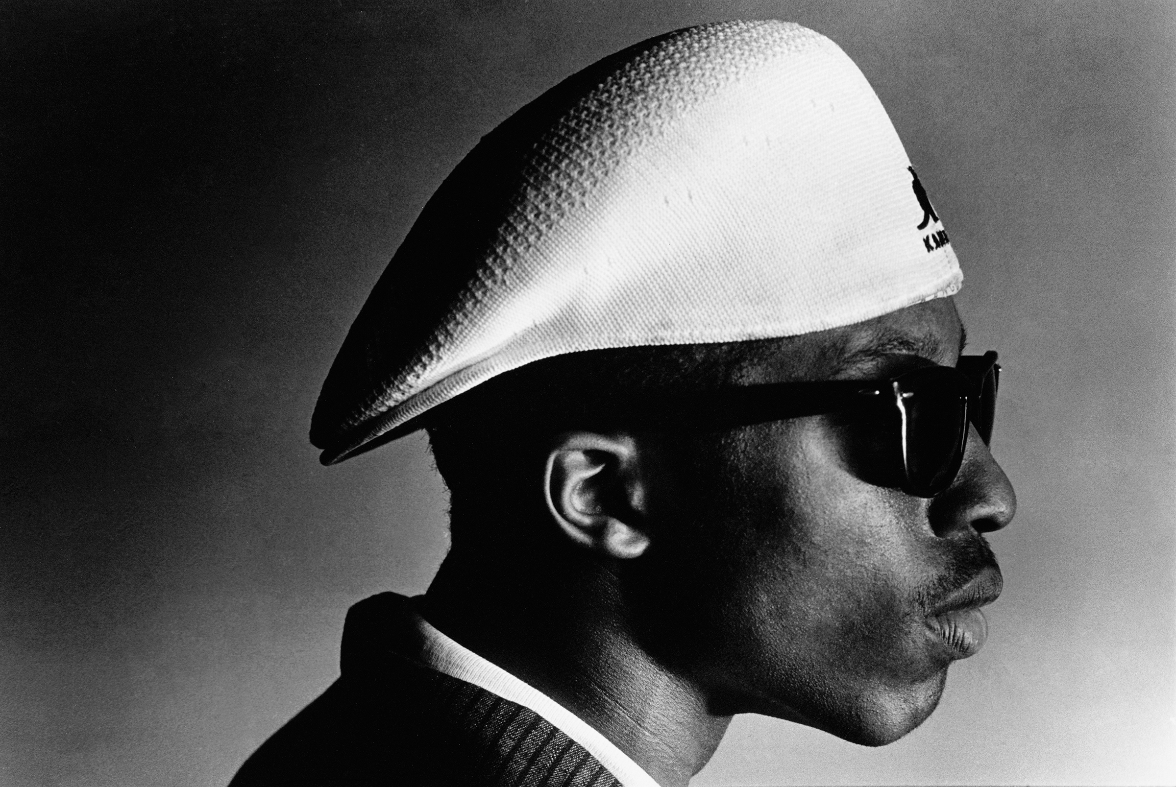 Black and white portrait showing man in profile wearing white beret and dark sunglasses against gradient grey background.