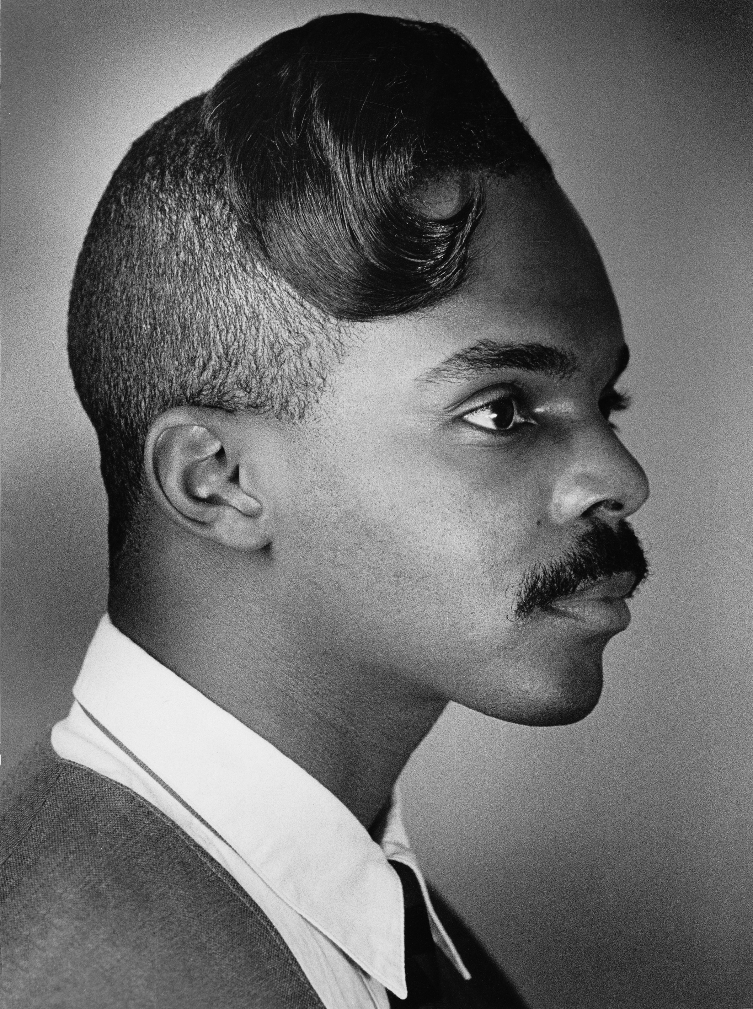 Black and white portrait of man in profile wearing white collar and tie, with distinctive rolled hairstyle and moustache.