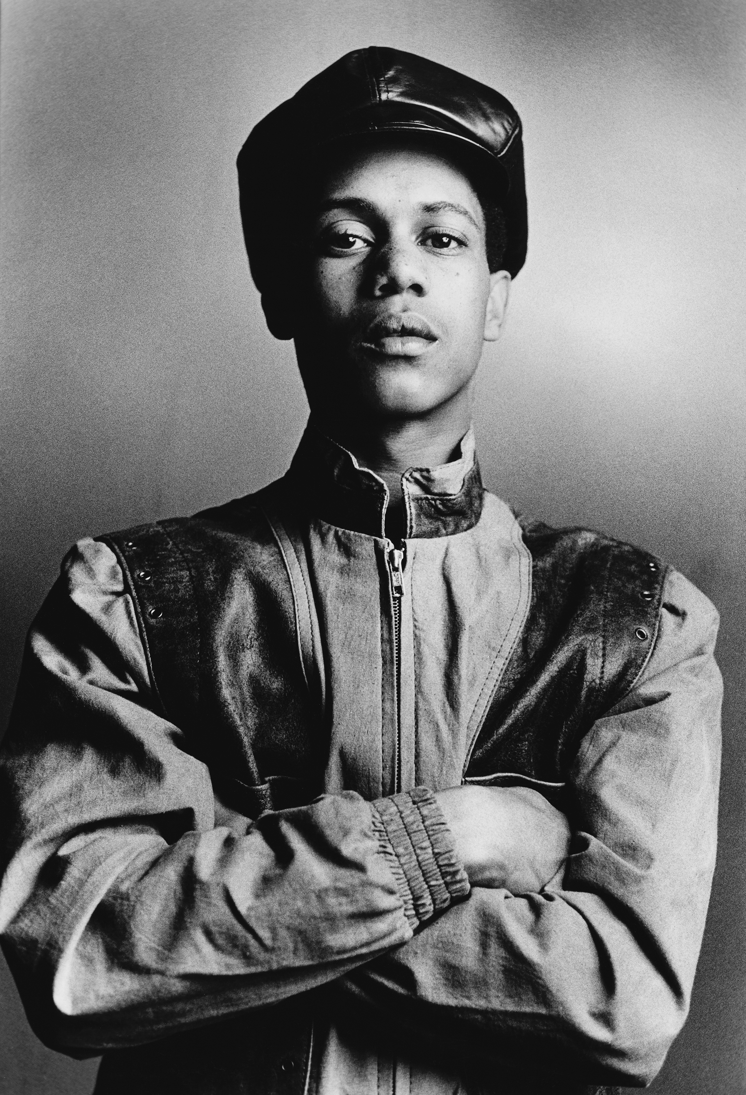 Black and white portrait of man with arms crossed wearing leather cap and zip-up leather jacket against plain background.