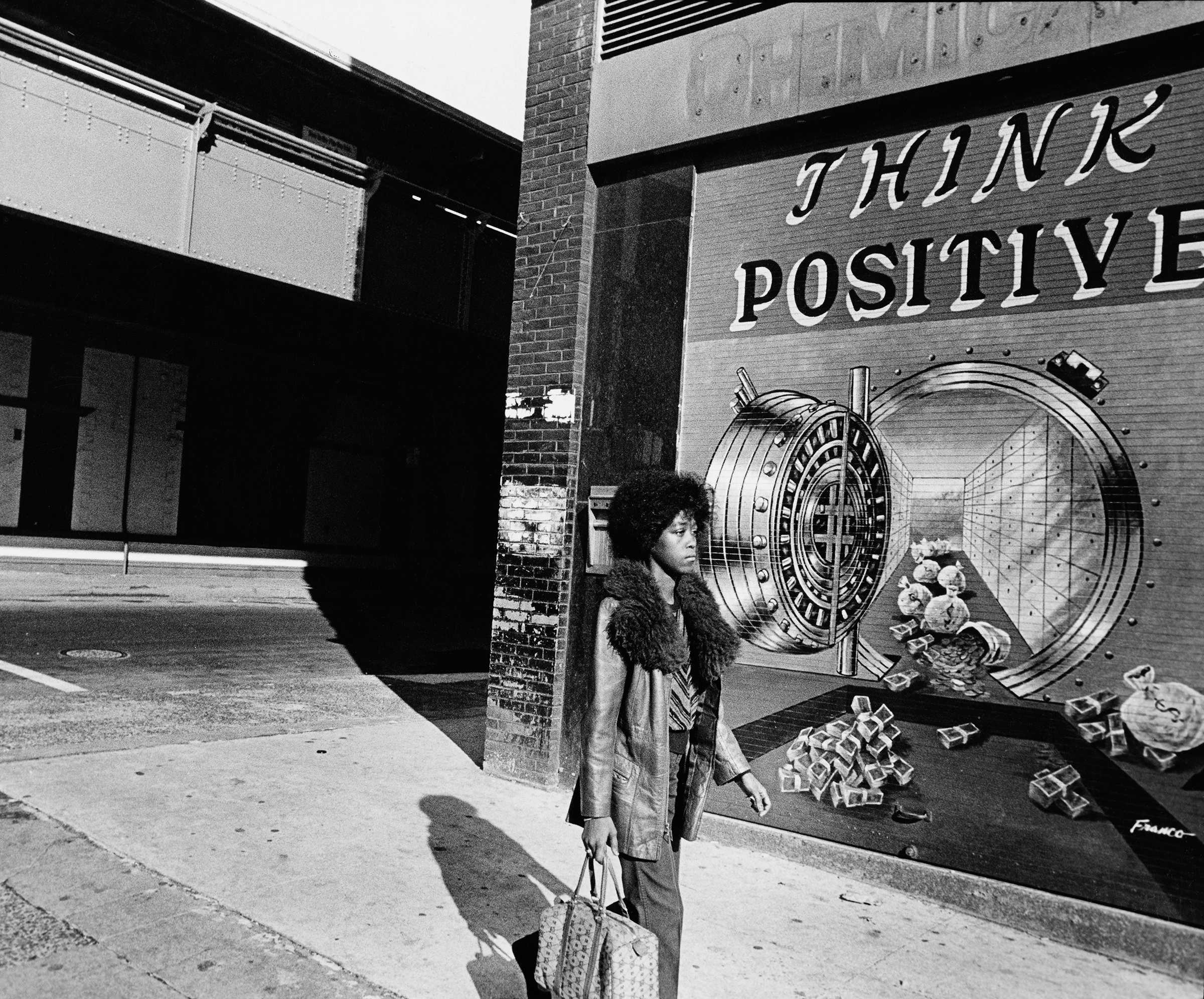 Black and white image showing a person standing beside a storefront with "THINK POSITIVE" text and large circular vault door graphic painted on the wall.