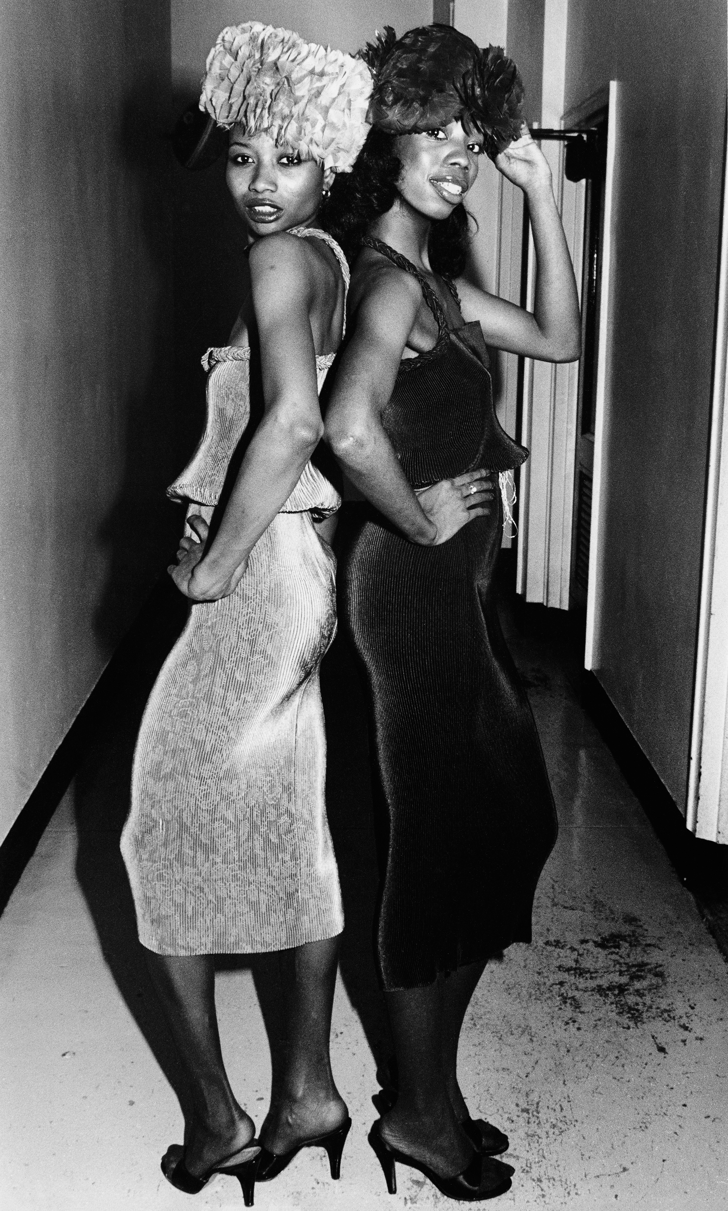 Black and white image of two women posing back-to-back in a doorway, wearing headscarves and dresses, one light-coloured, one dark.