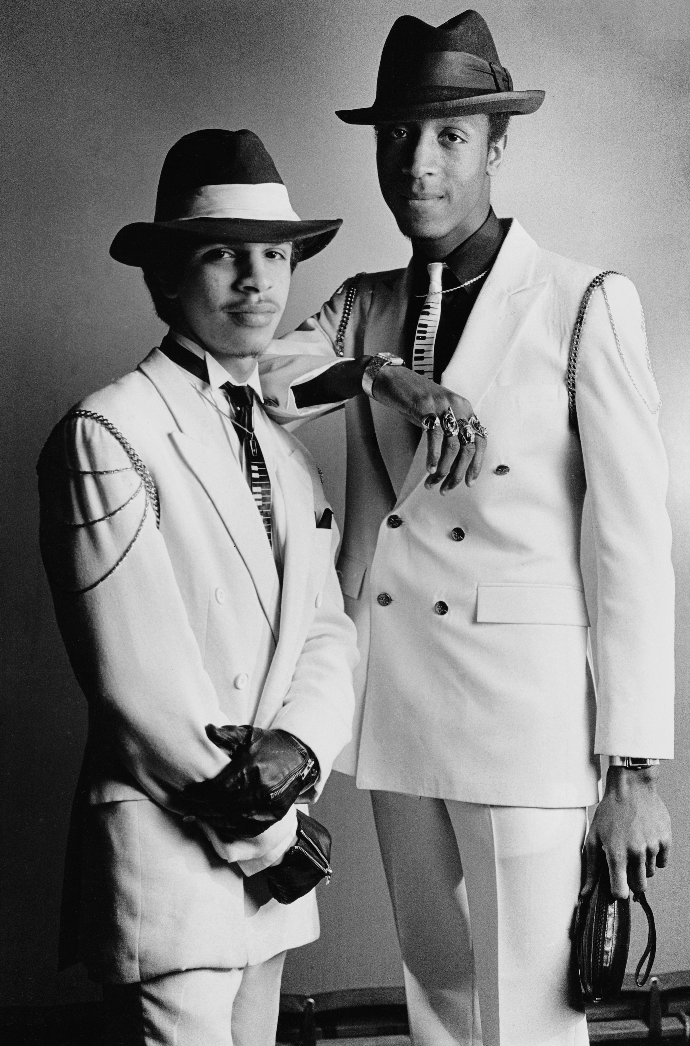 Black and white portrait of two men in white suits and dark fedoras, one holding a saxophone, against a plain background.