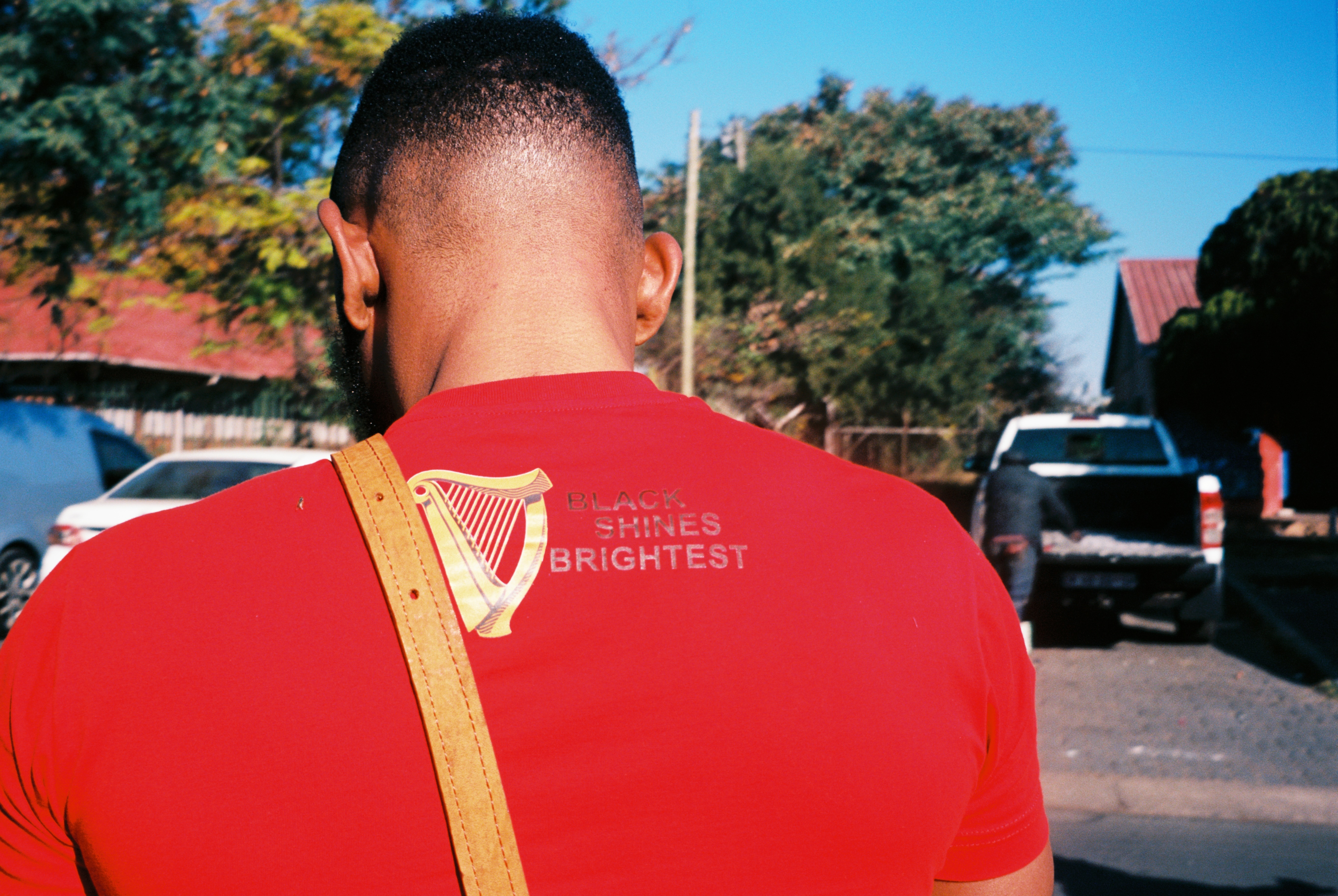 A person wearing a bright red shirt with the text "She's brightest" printed on the back, standing in front of a row of trees and buildings.