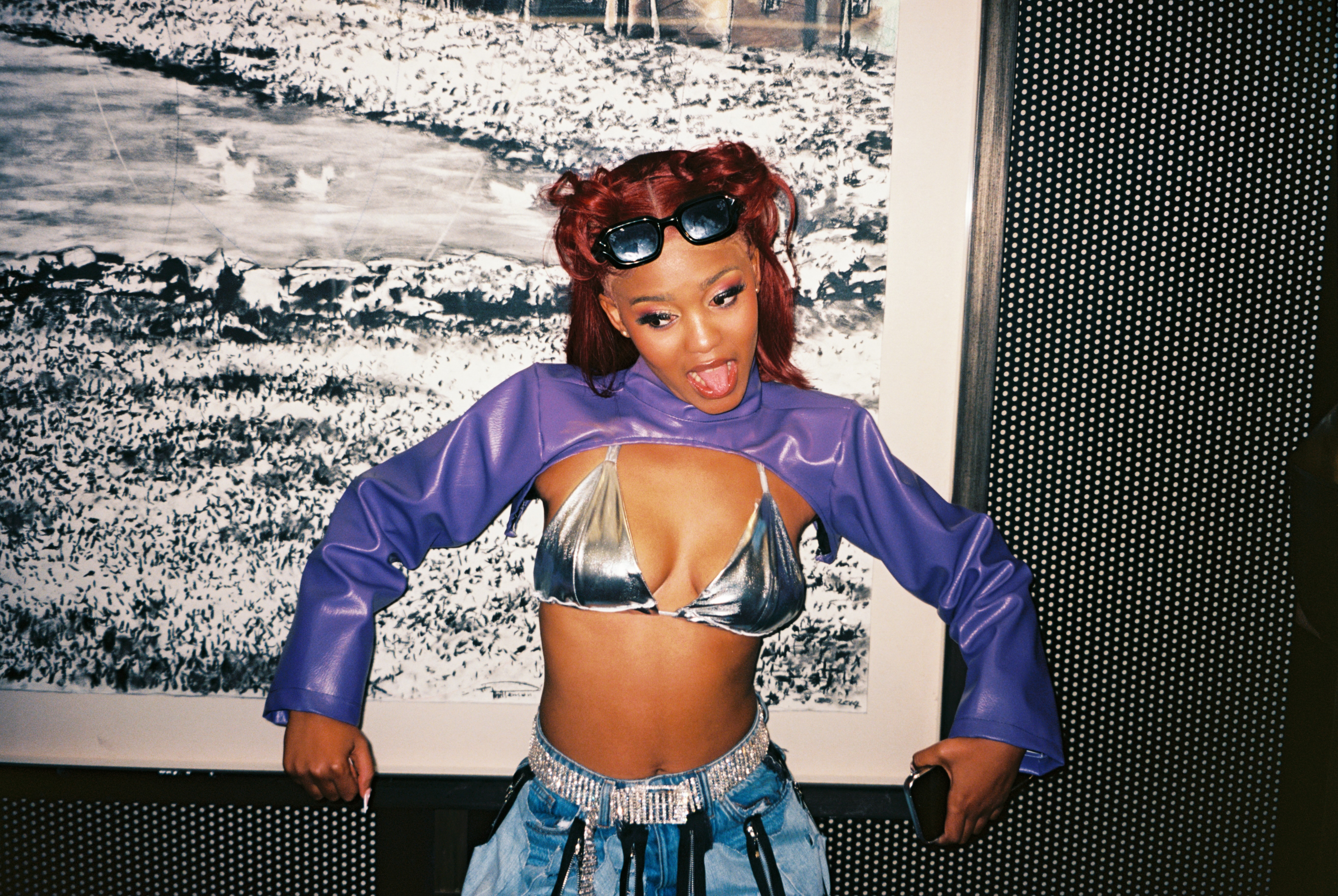 A smiling woman with red curly hair wearing a purple cropped top, sunglasses, and distressed jeans, standing in front of a blurred outdoor scene.