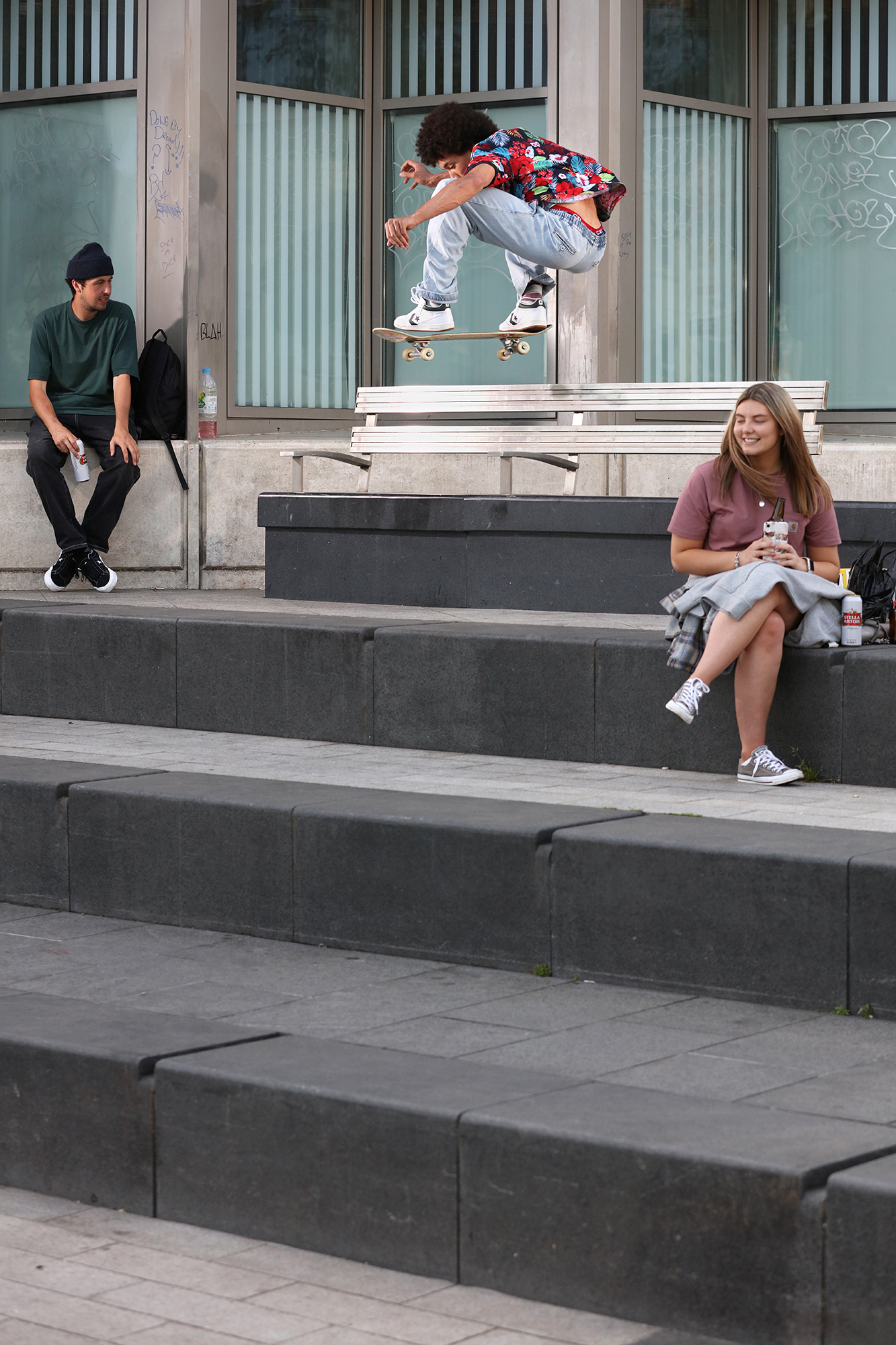 Skateboarder jumping over concrete steps whilst two people sit nearby watching outside modern building with glass windows.