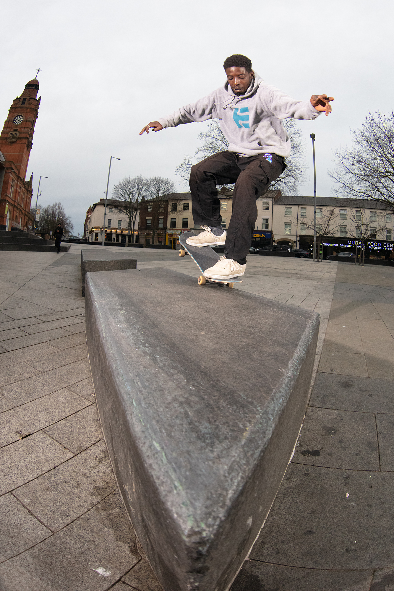 Skateboarder in grey hoodie performing trick on concrete ledge at urban skate park with Victorian buildings in background.