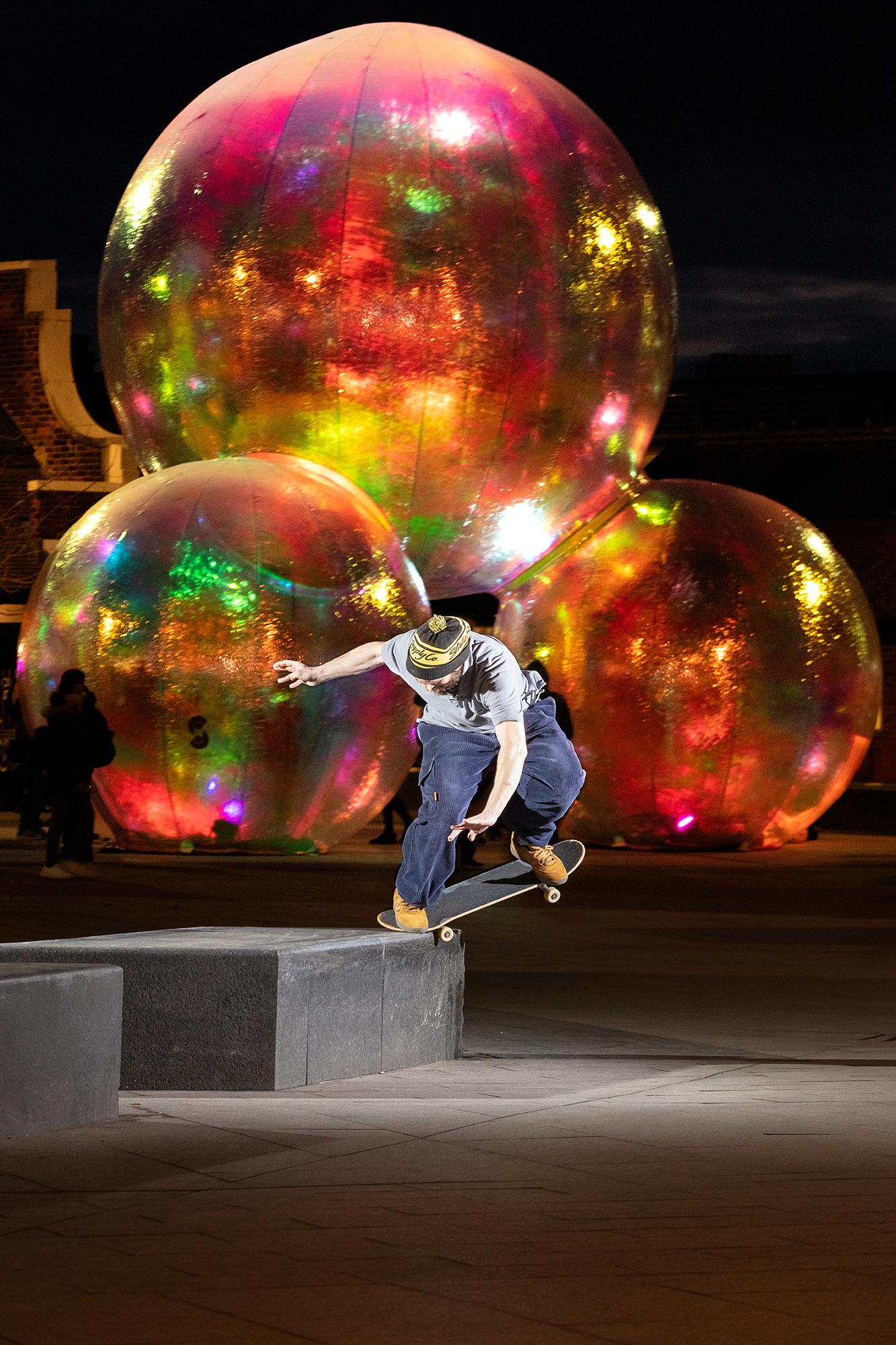 Skateboarder jumping over concrete ledge with large iridescent spheres displaying rainbow colours in background at night.