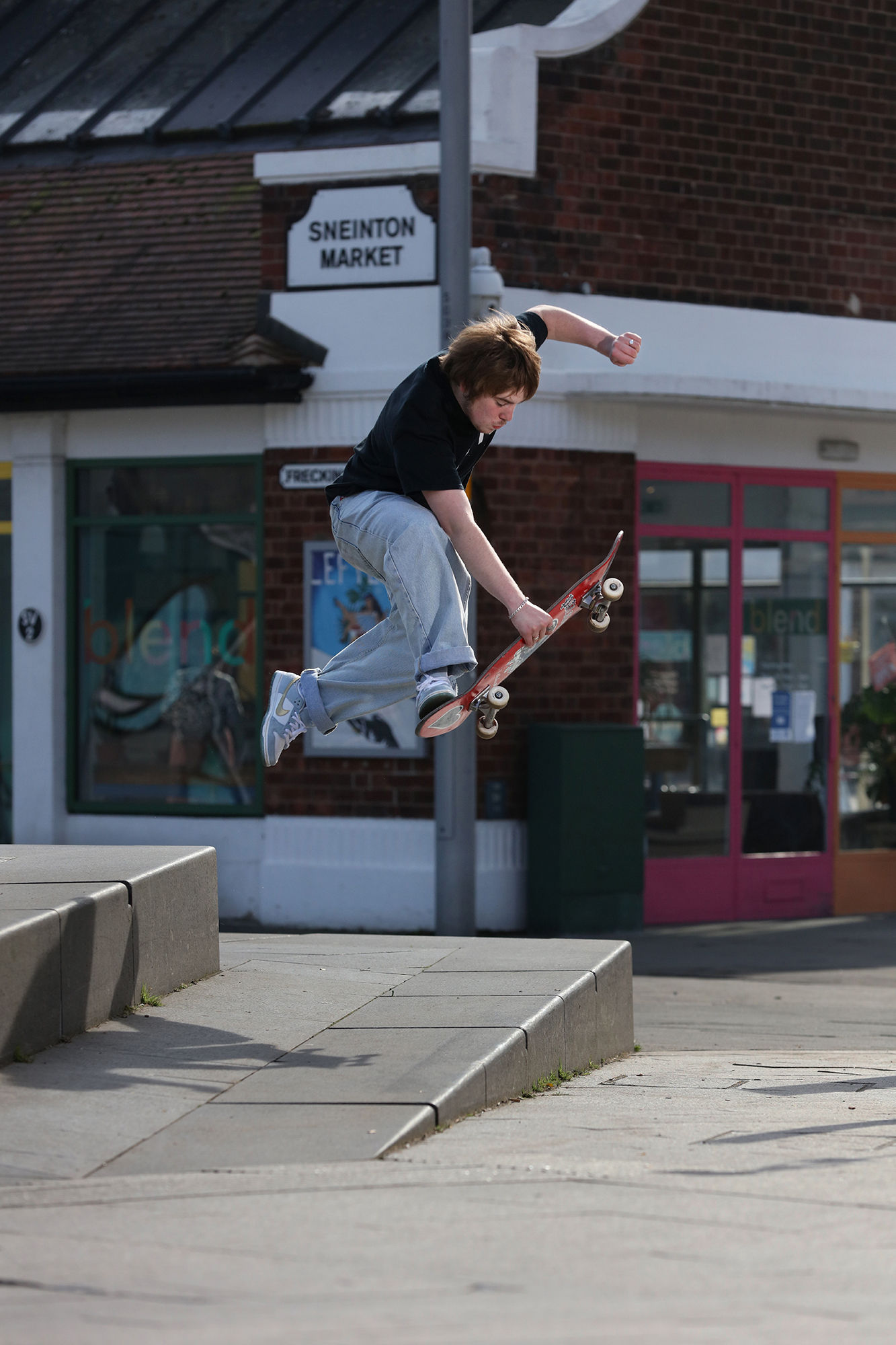 Skateboarder performing aerial trick over concrete ledge outside Sneinton Market building with brick facade and shop fronts.