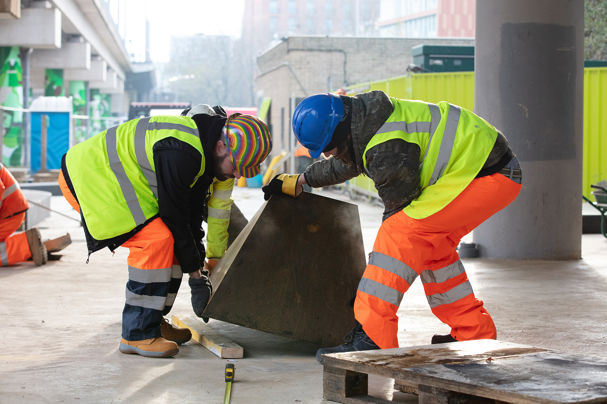 Two construction workers in high-vis jackets and hard hats examining concrete block on building site with concrete pillars in background.