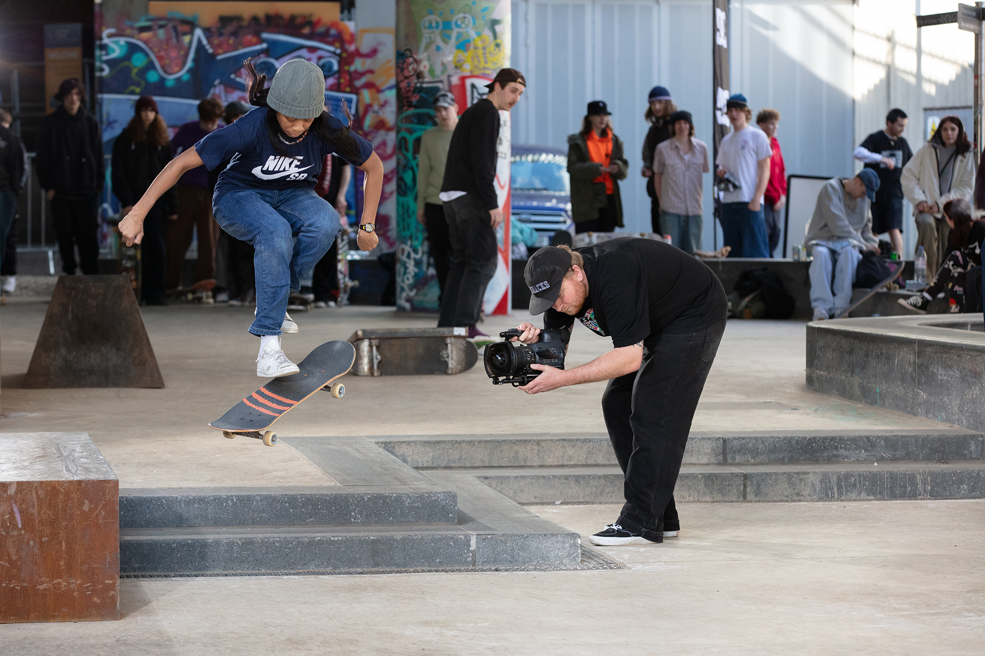 Skateboarders at concrete skate park with ramps and steps, spectators watching, colourful graffiti on walls in background.