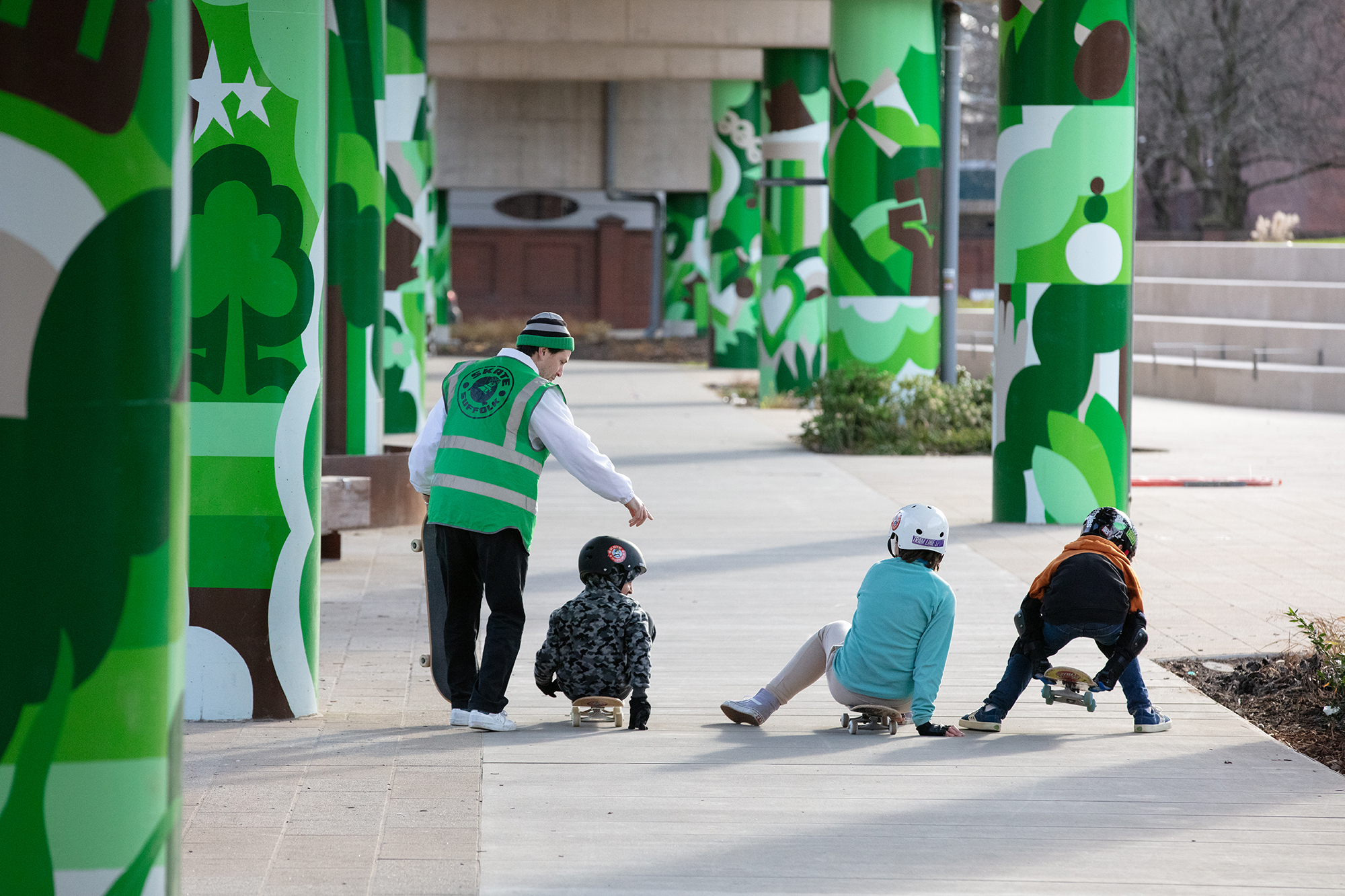 Adult in green hi-vis vest with four children on concrete plaza surrounded by green geometric tree murals on columns.