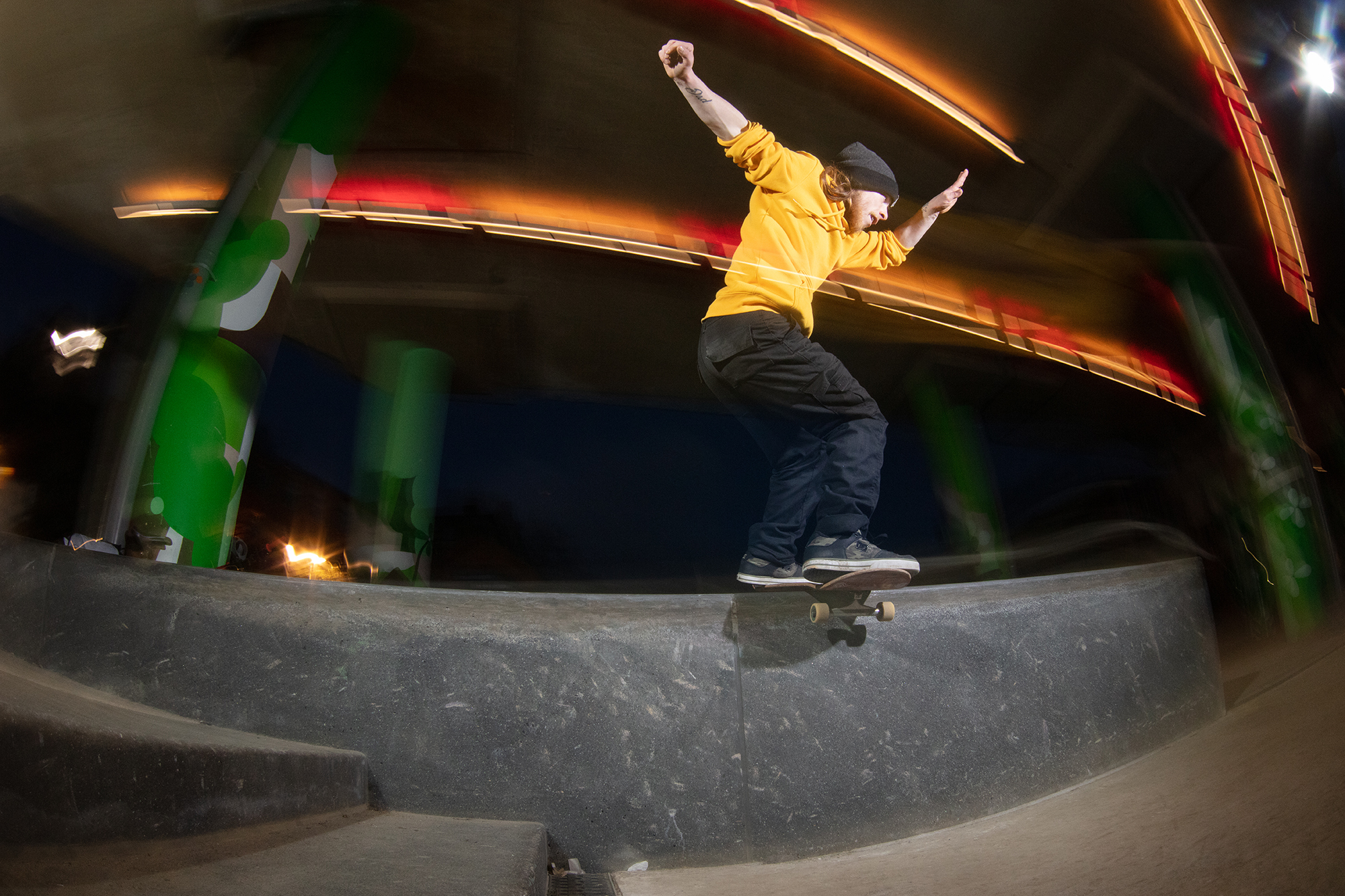 Skateboarder in yellow jumper mid-air above curved concrete bowl, with blurred coloured lights and figures in background.