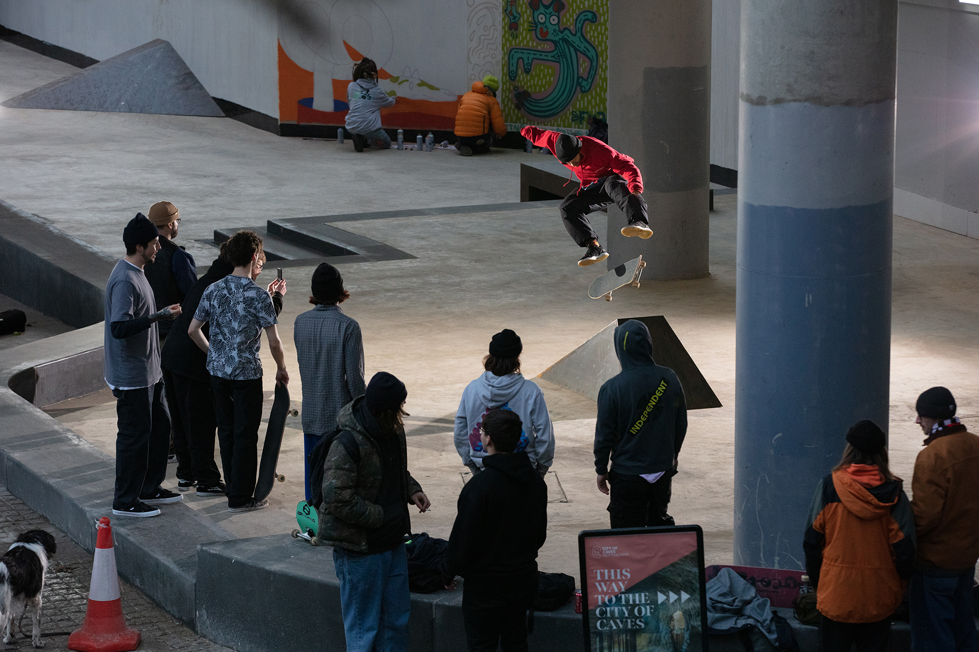 Skateboarder in red performing trick on concrete ramp whilst group of people watch from below in indoor skate park.