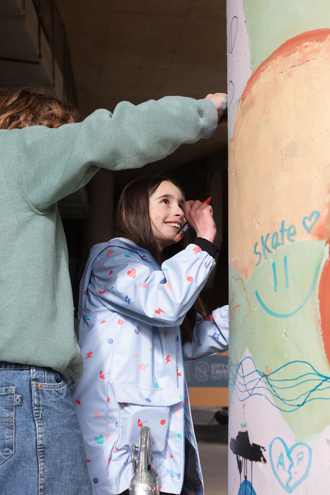 Two people in casual clothing standing by a wall with colourful graffiti and chalk drawings, one wearing patterned pyjamas.