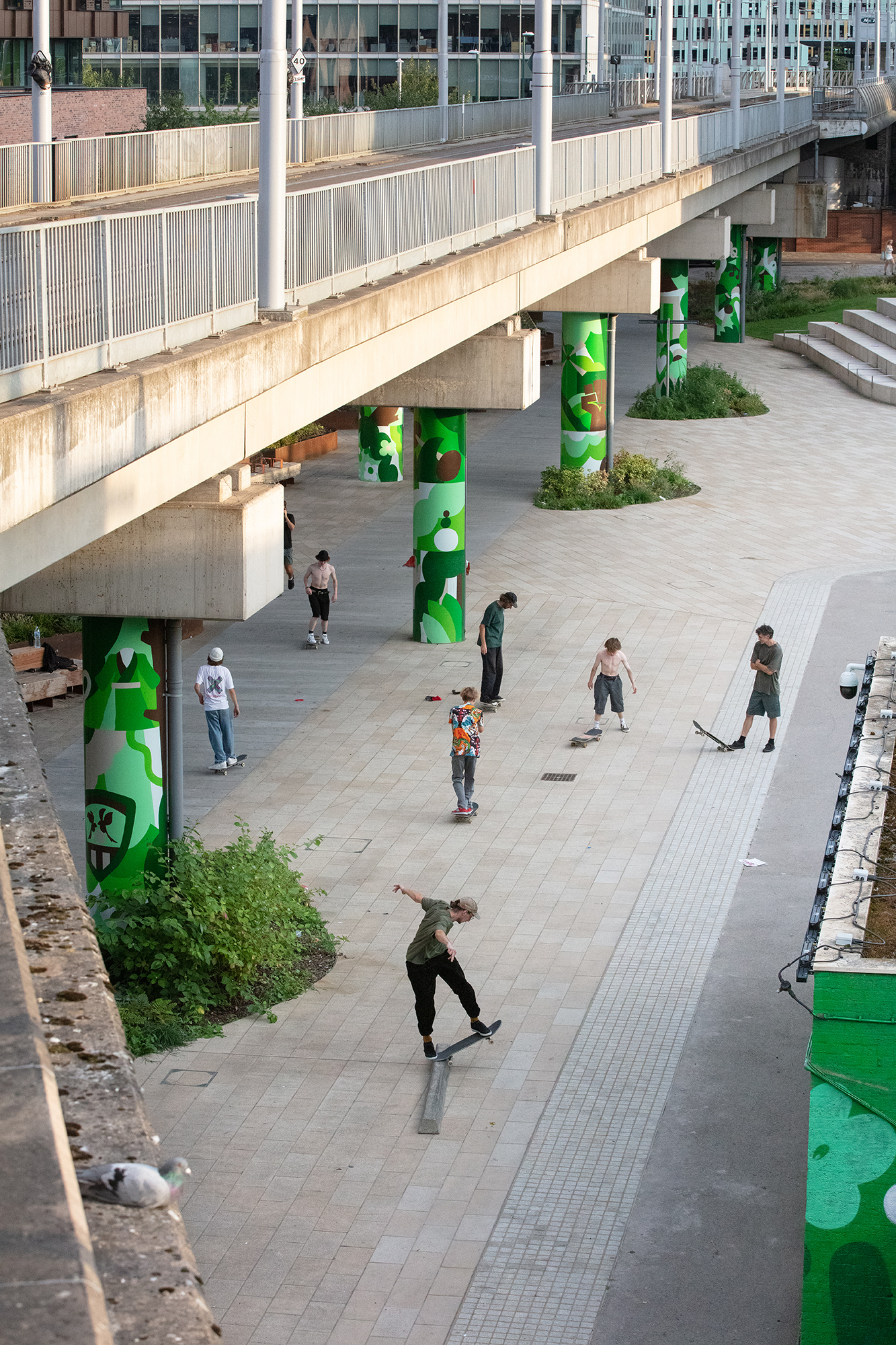 Concrete pedestrian bridge with white railings spans plaza where people skateboard. Green-painted support pillars, grey paving stones, modern buildings in background.