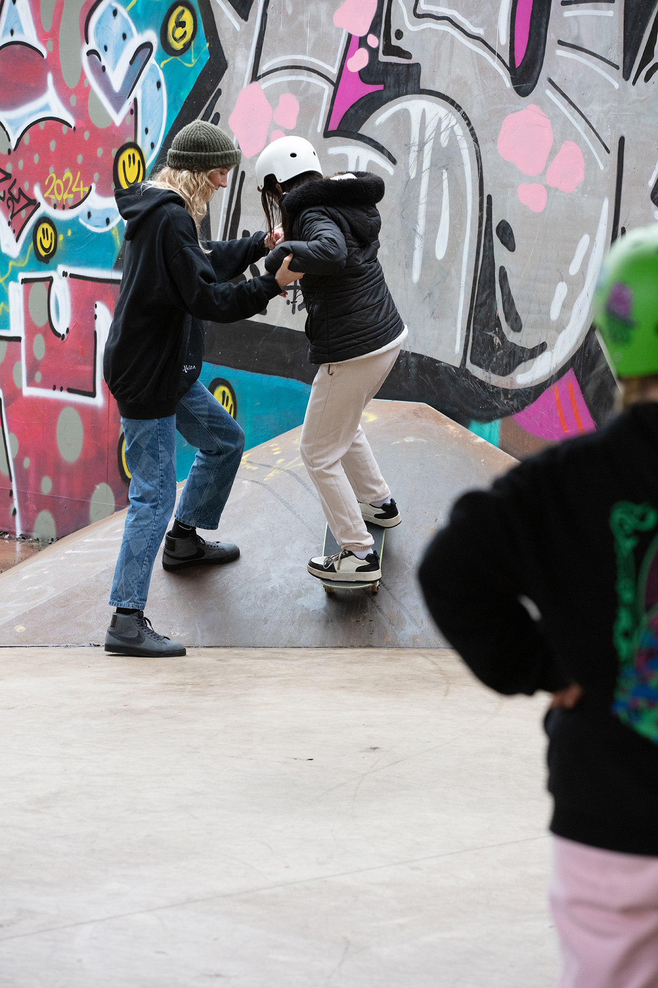 Two people in dark clothing and hoodies spray painting colourful graffiti on concrete wall with pink, blue, and yellow designs.