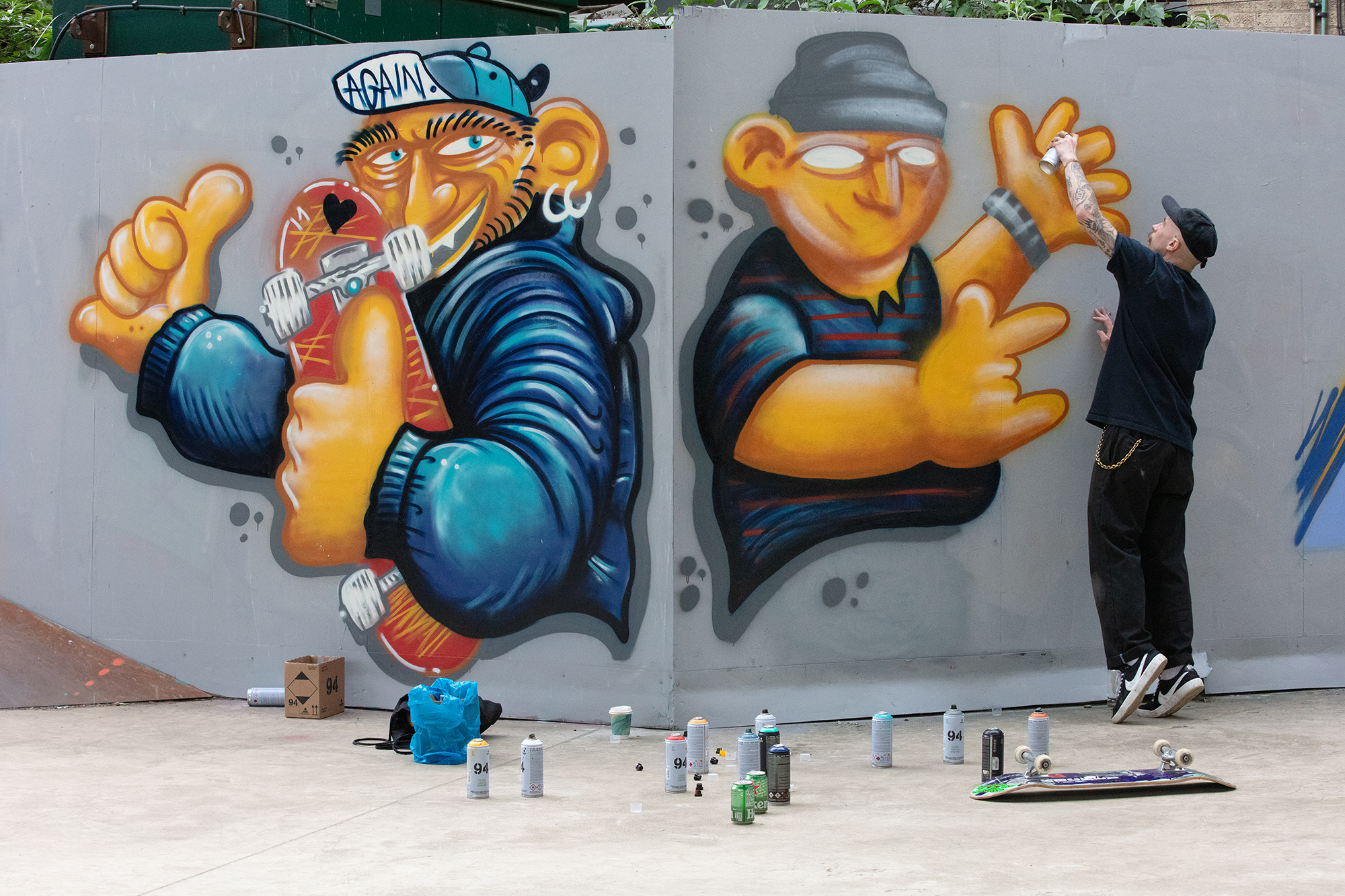 Street artist painting mural on white wall featuring two cartoon characters in blue clothing with orange skin and exaggerated features.
