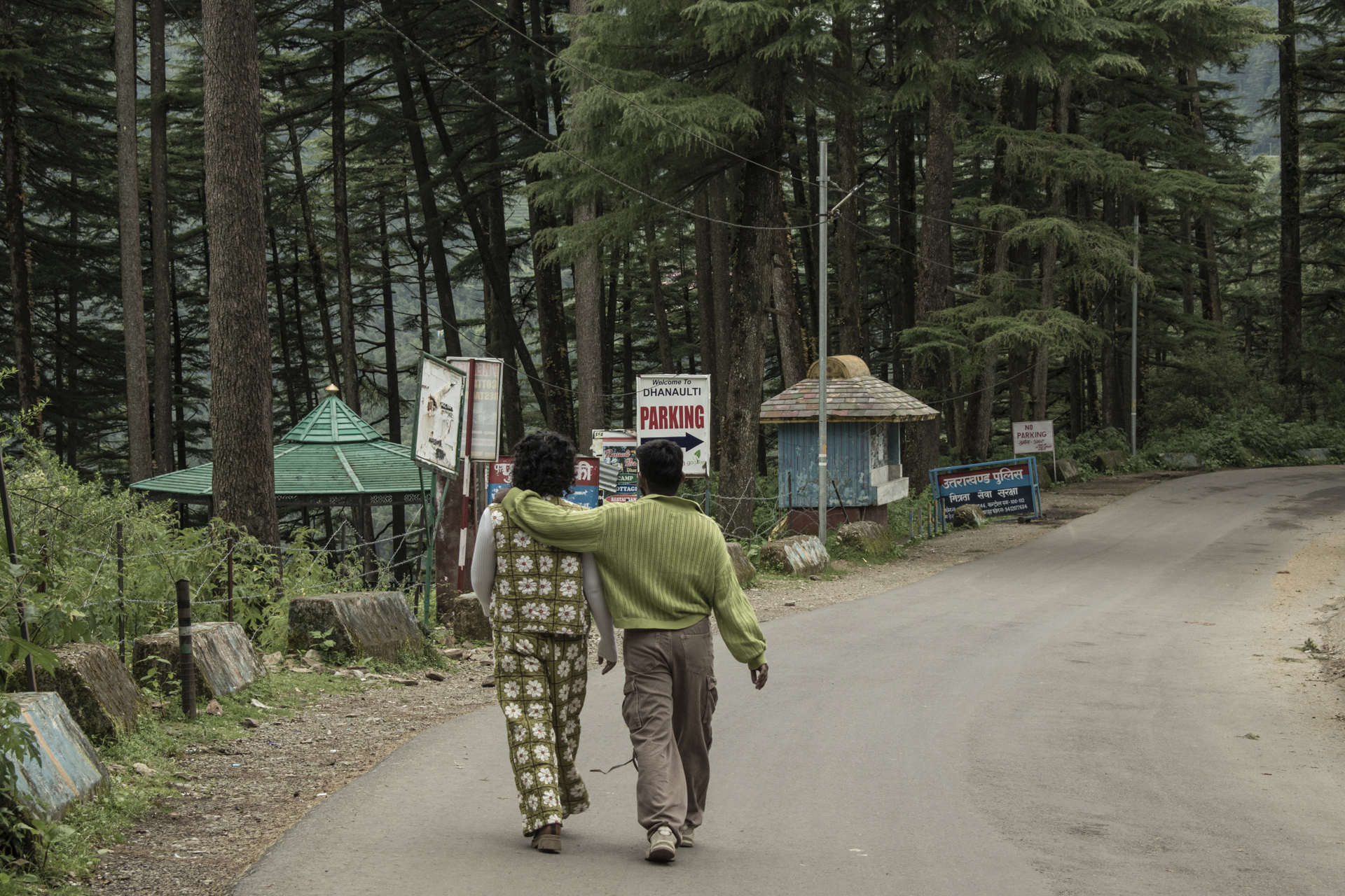 Person in green jacket carrying large basket walks along dirt road past small roadside stalls surrounded by dense forest.