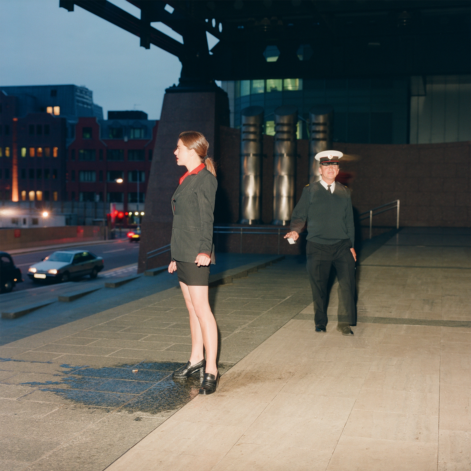 Woman in green coat and man in naval uniform standing on rooftop at dusk, city buildings with lit windows in background.