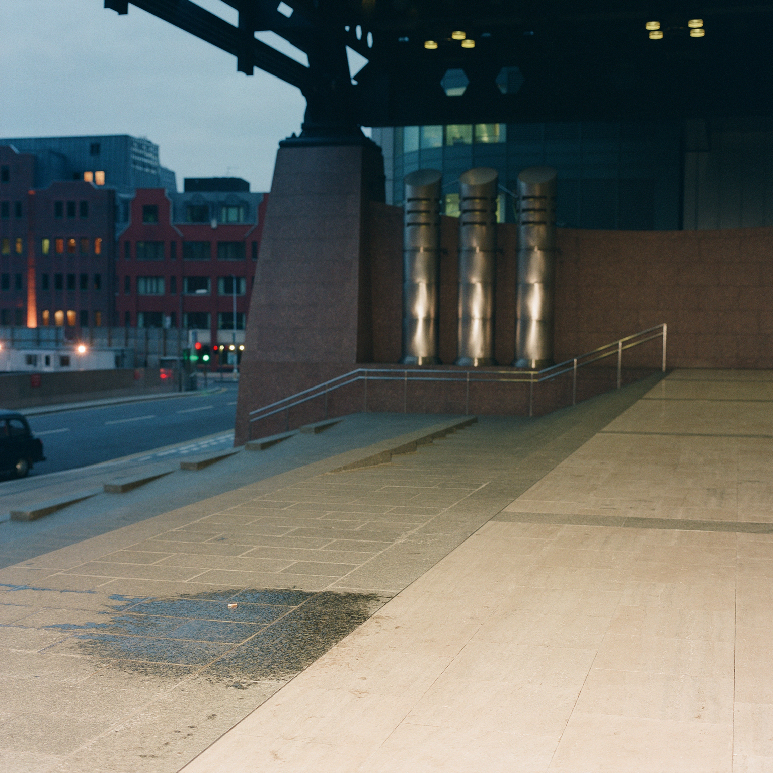 Urban evening scene with illuminated cylindrical columns against concrete wall, paved walkway, and city buildings with lit windows in background.