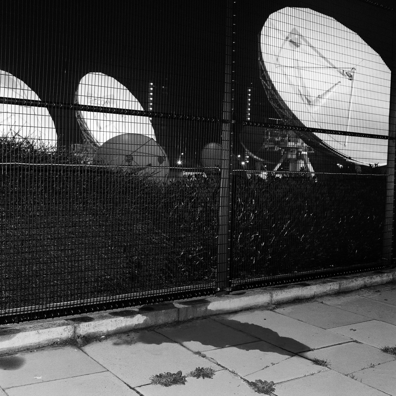 Large satellite dishes behind wire mesh fencing, viewed through curved architectural openings in black and white.