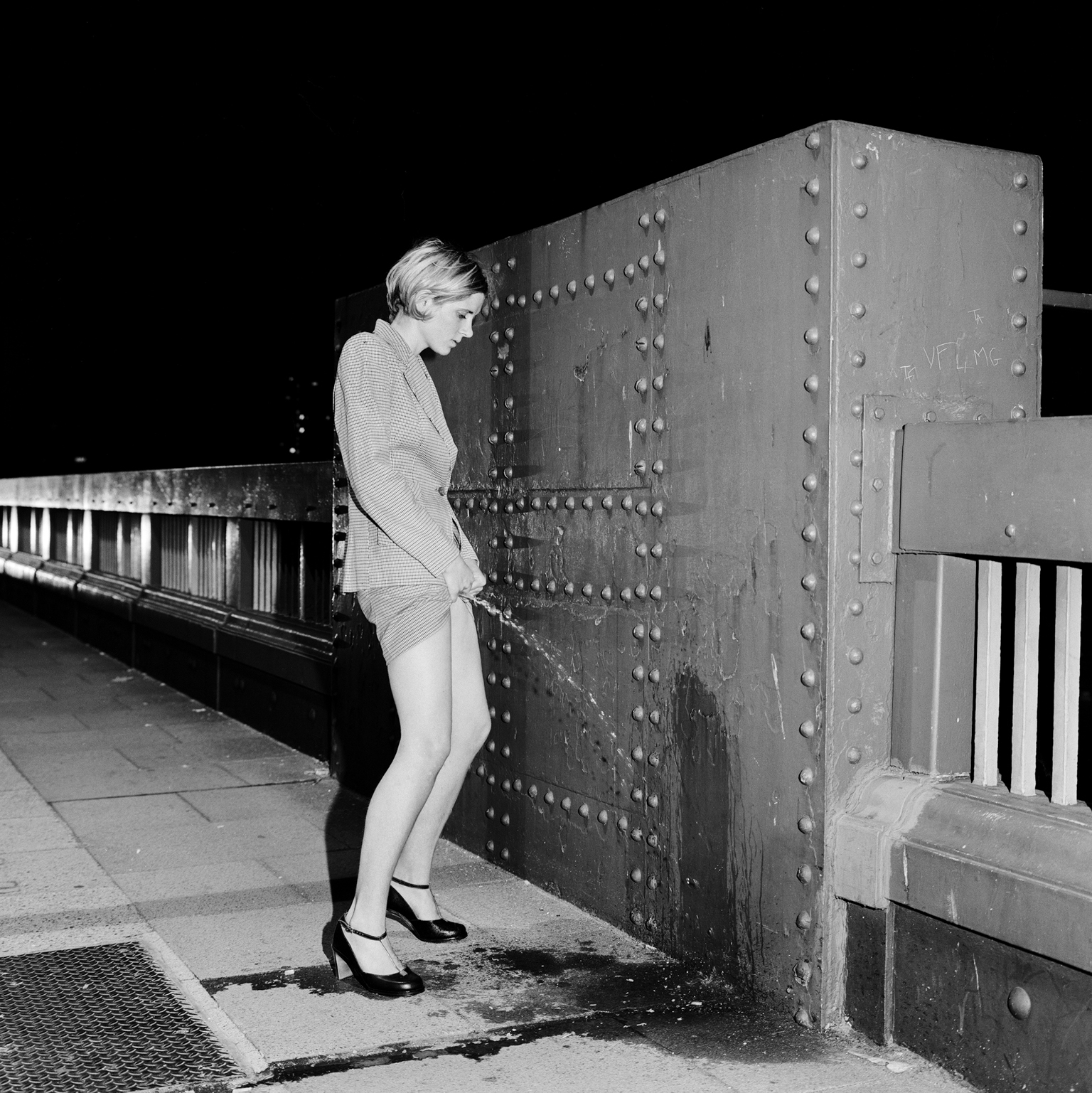 Woman in light-coloured dress leaning against large riveted metal bridge structure with railings at night, black and white photograph.