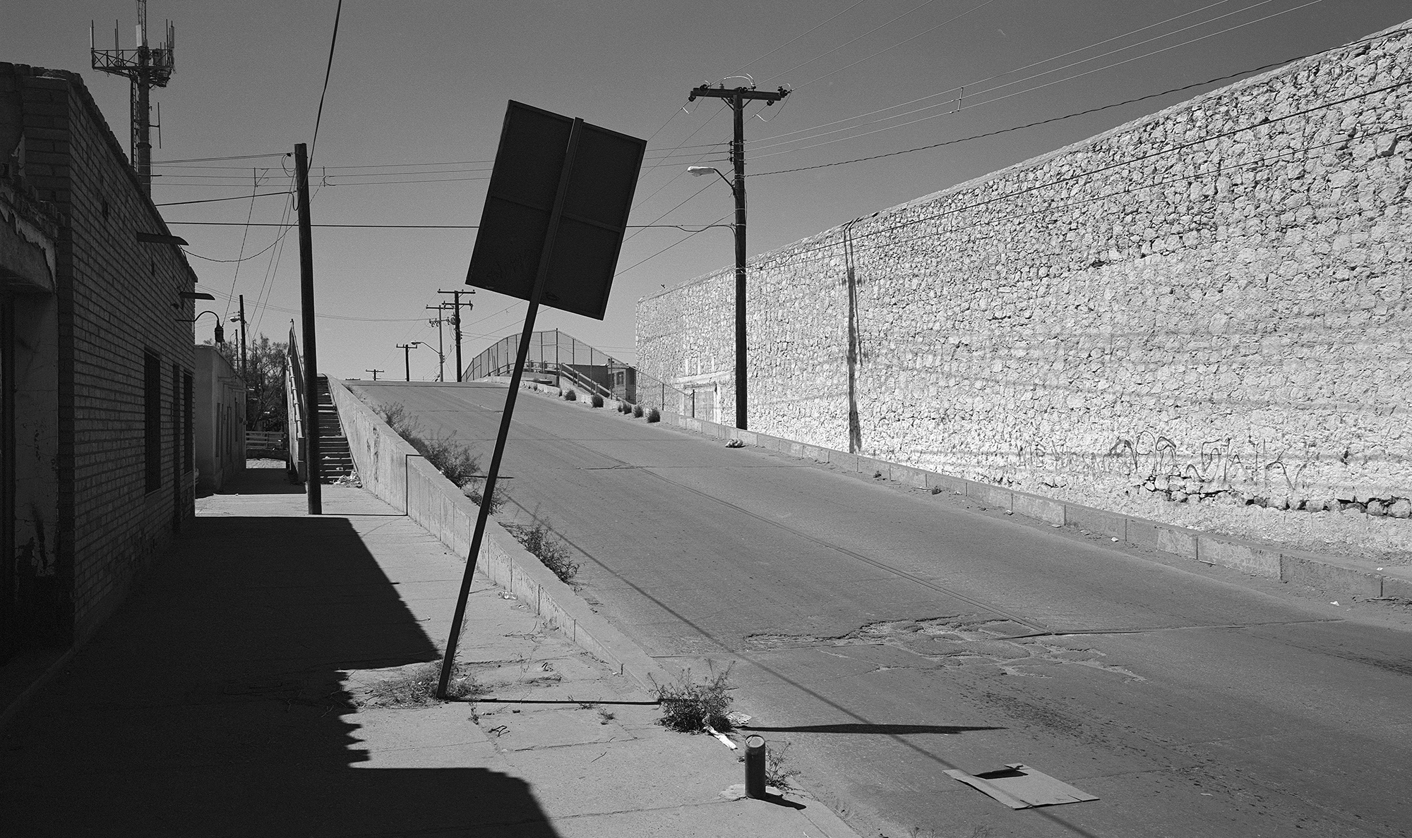 Black and white image showing tilted road sign on empty street with telephone poles, buildings on left, and hillside on right.