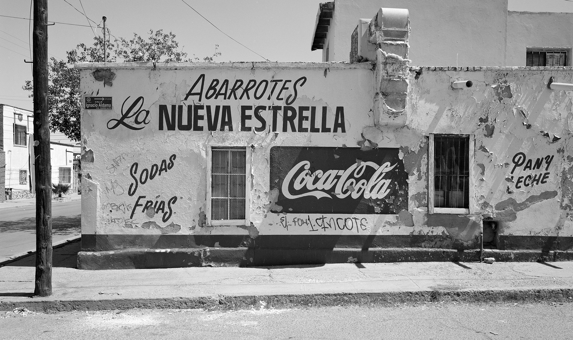 Black and white image of weathered building wall with Spanish text "ABARROTES La NUEVA ESTRELLA", "Sopas Frias", and Coca-Cola signage painted on facade.