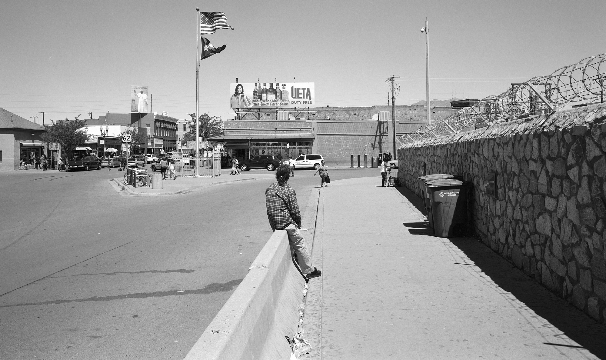 Man sweeping street with broom in black and white image. Stone wall right side, buildings and flagpole in background.