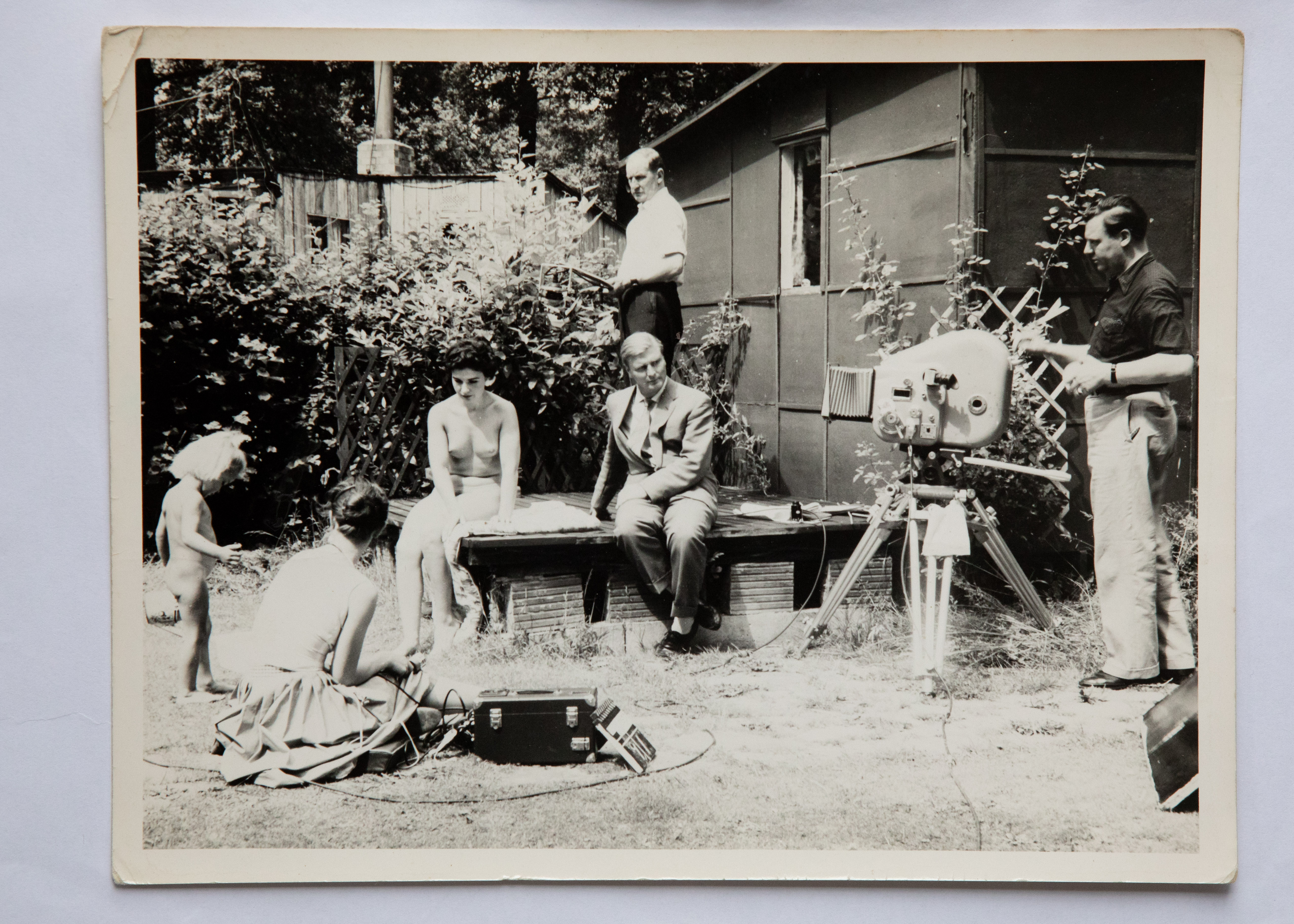 Black and white vintage photograph showing people gathered around wooden building with garden tools and equipment scattered about.