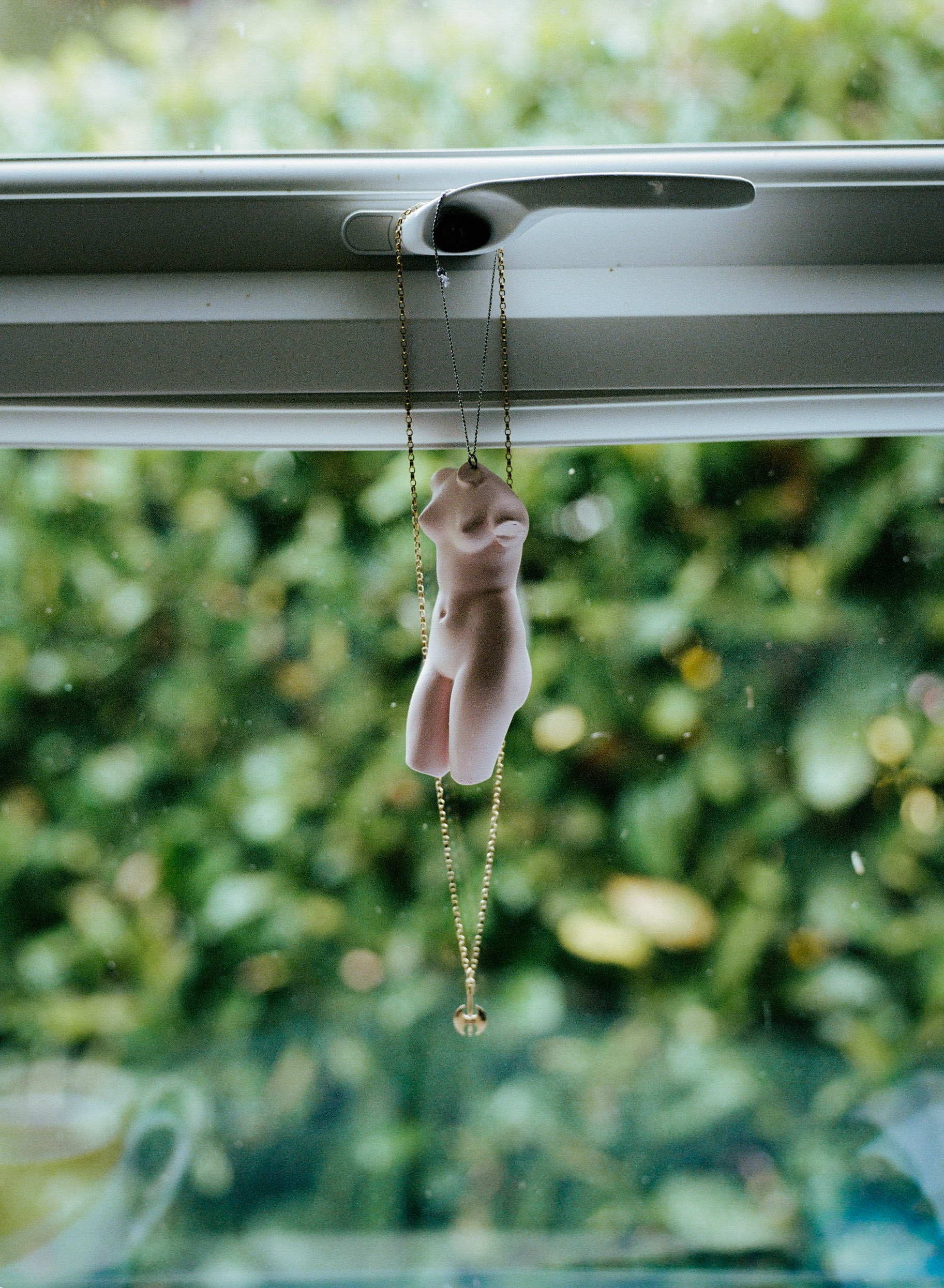 Miniature plastic hand hanging from chain attached to window latch, with blurred green foliage visible outside.