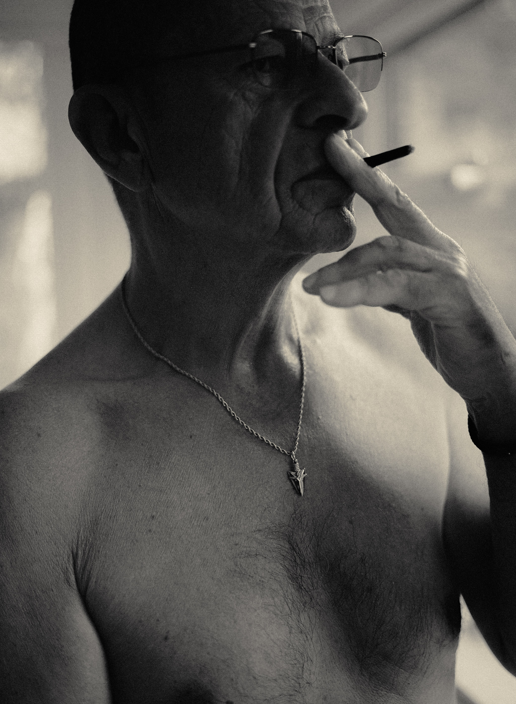 Shirtless man with glasses and necklace smoking cigarette, hand raised to mouth, black and white portrait.
