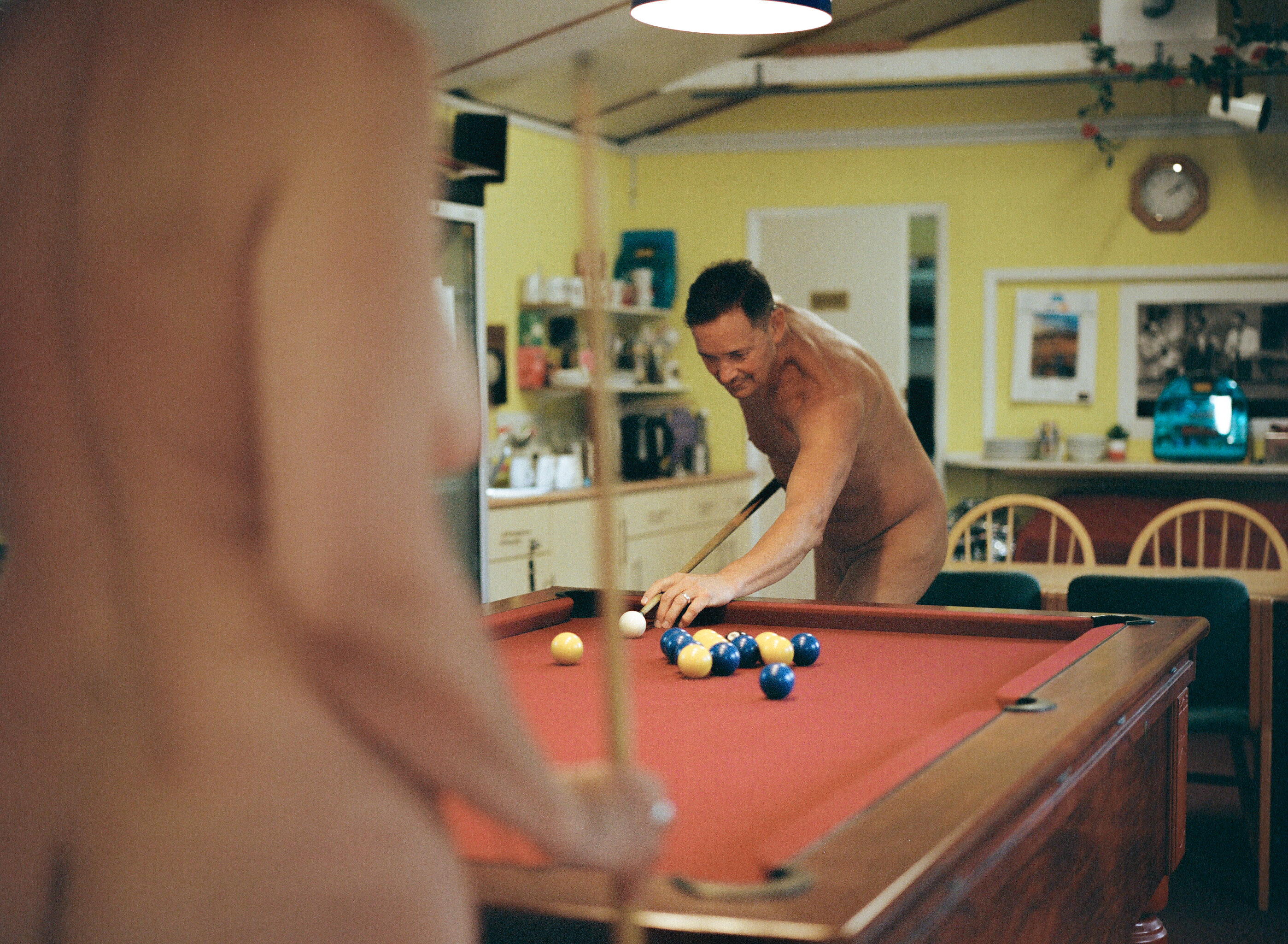 Shirtless man playing pool on red felt table in yellow kitchen, viewed from behind another person's shoulder in foreground.