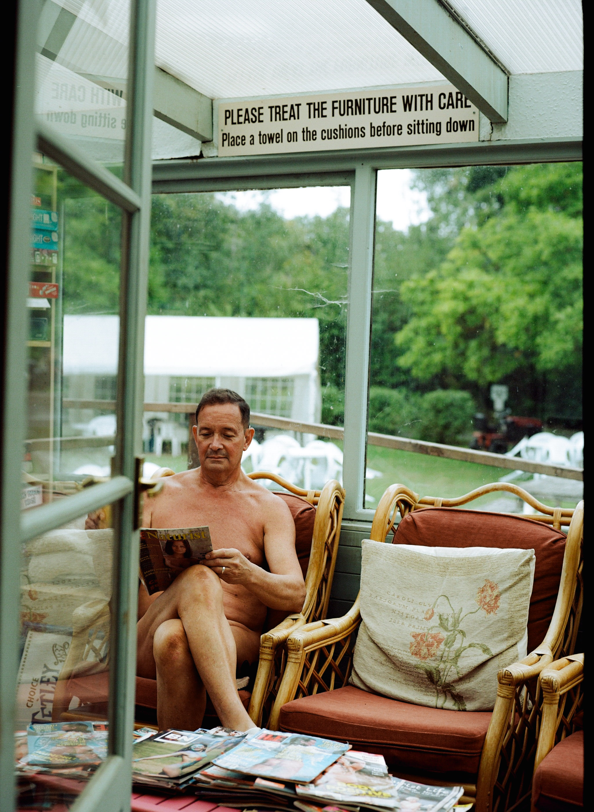 Shirtless man sitting in wooden chair inside glass conservatory with sign about treating furniture carefully, green garden visible outside.