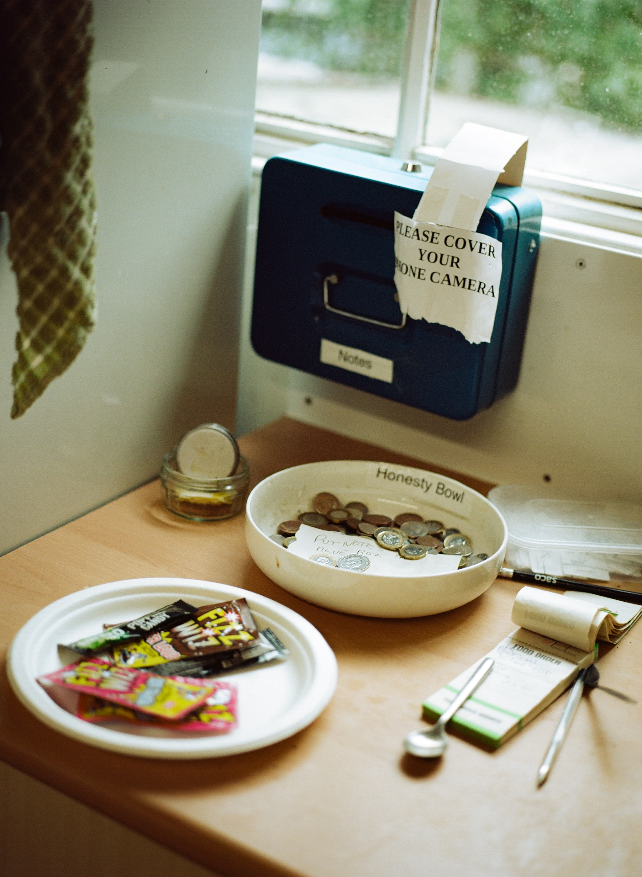 Black Polaroid camera on windowsill with white note reading "PLEASE COVER YOUR PHONE CAMERA". Two white bowls below contain coins and sweets.