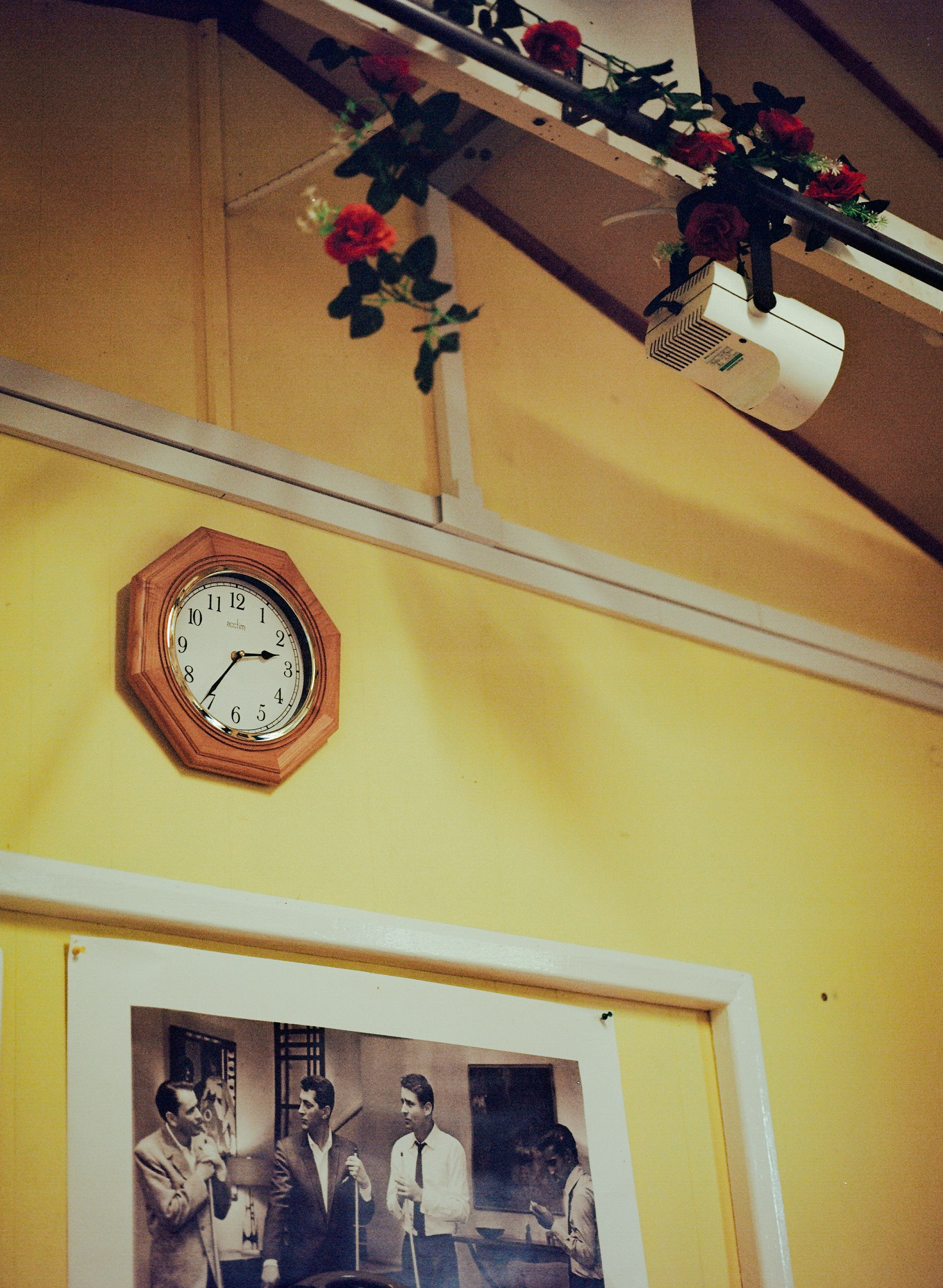 Yellow wall with octagonal wooden clock, framed black-and-white photograph, and exposed beam with red roses hanging above.