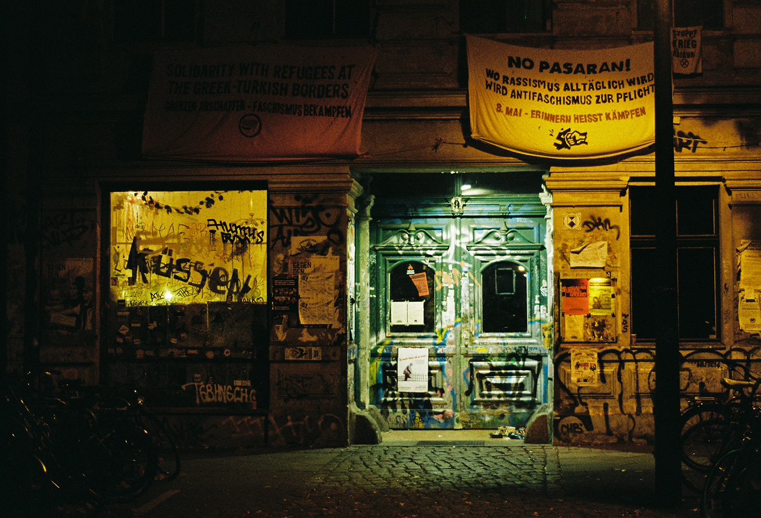 Rundown urban building façade at night, with graffiti-covered walls, signs, and illuminated storefront windows.