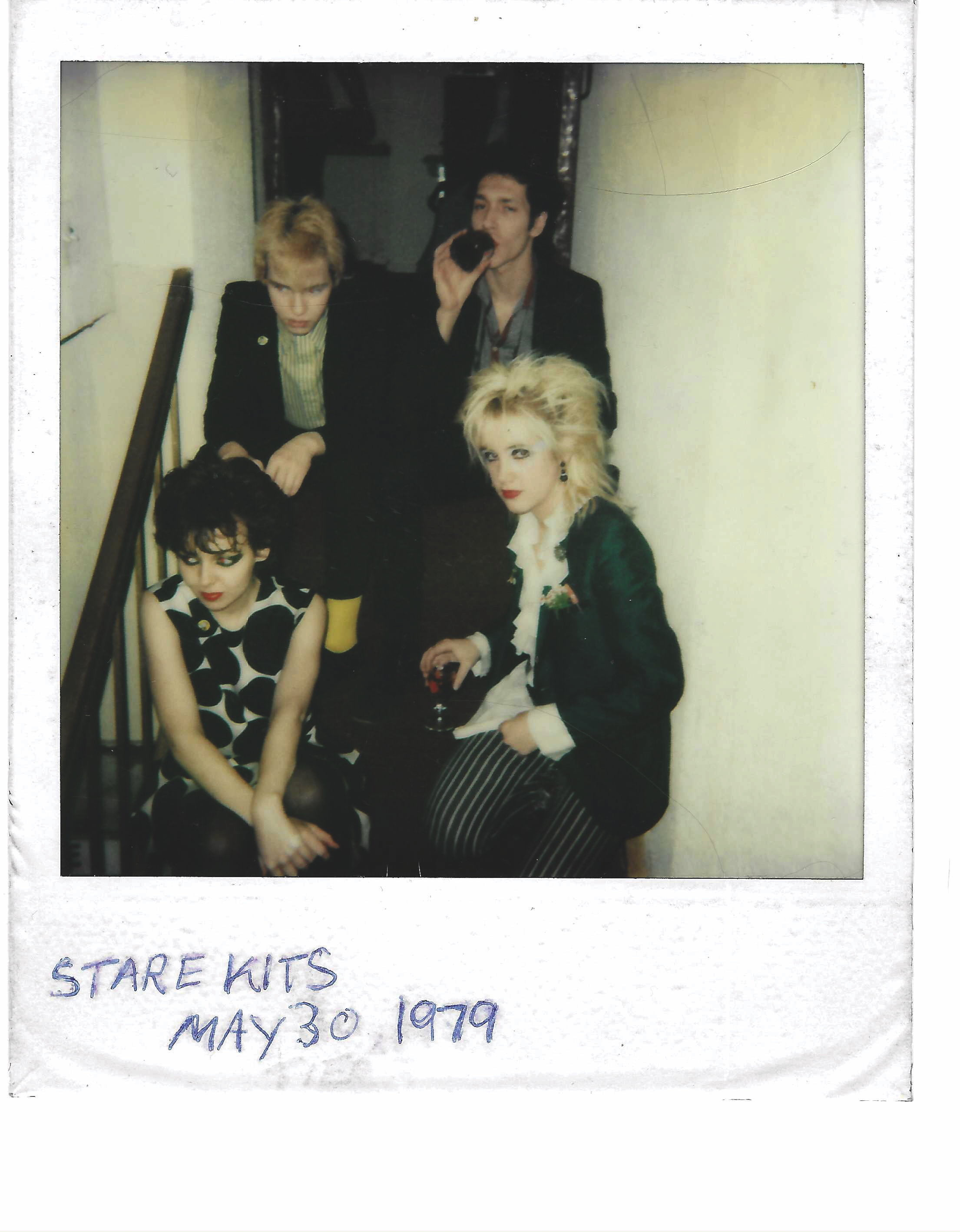 A group of four people in dark clothing, posing together in a theatre or venue setting. The image has a vintage, polaroid-style appearance.