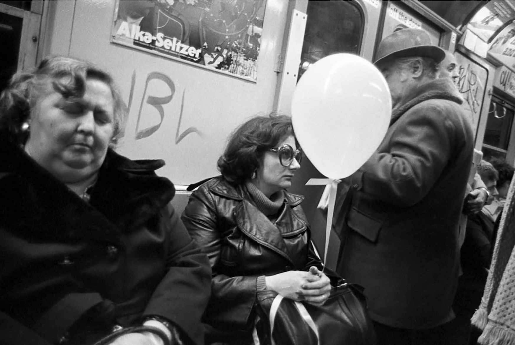 Monochrome image of people sitting on a train carriage, one person holding a white balloon.