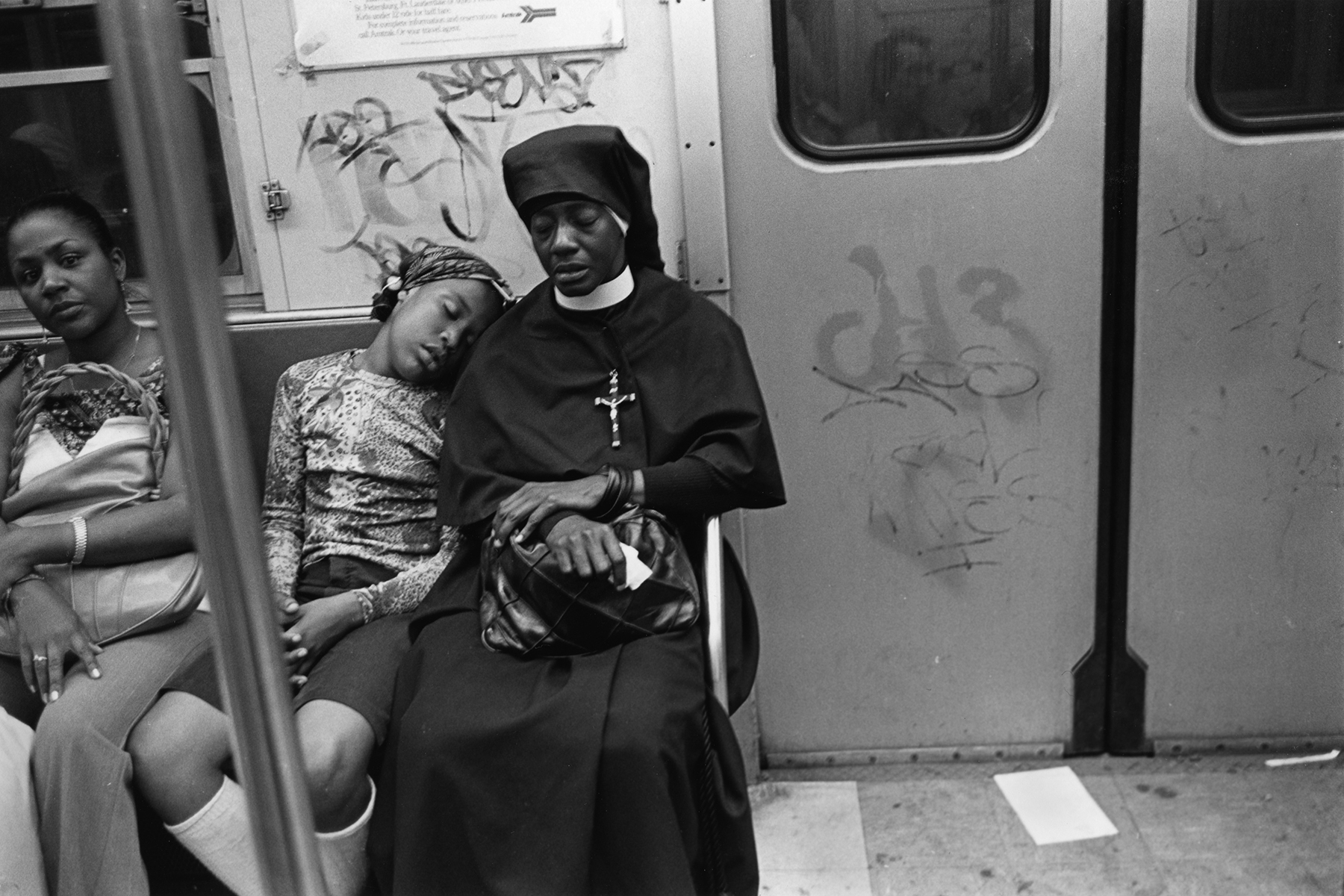 A black-and-white image showing a nun embracing a woman on a subway carriage. Graffiti can be seen on the walls behind them.