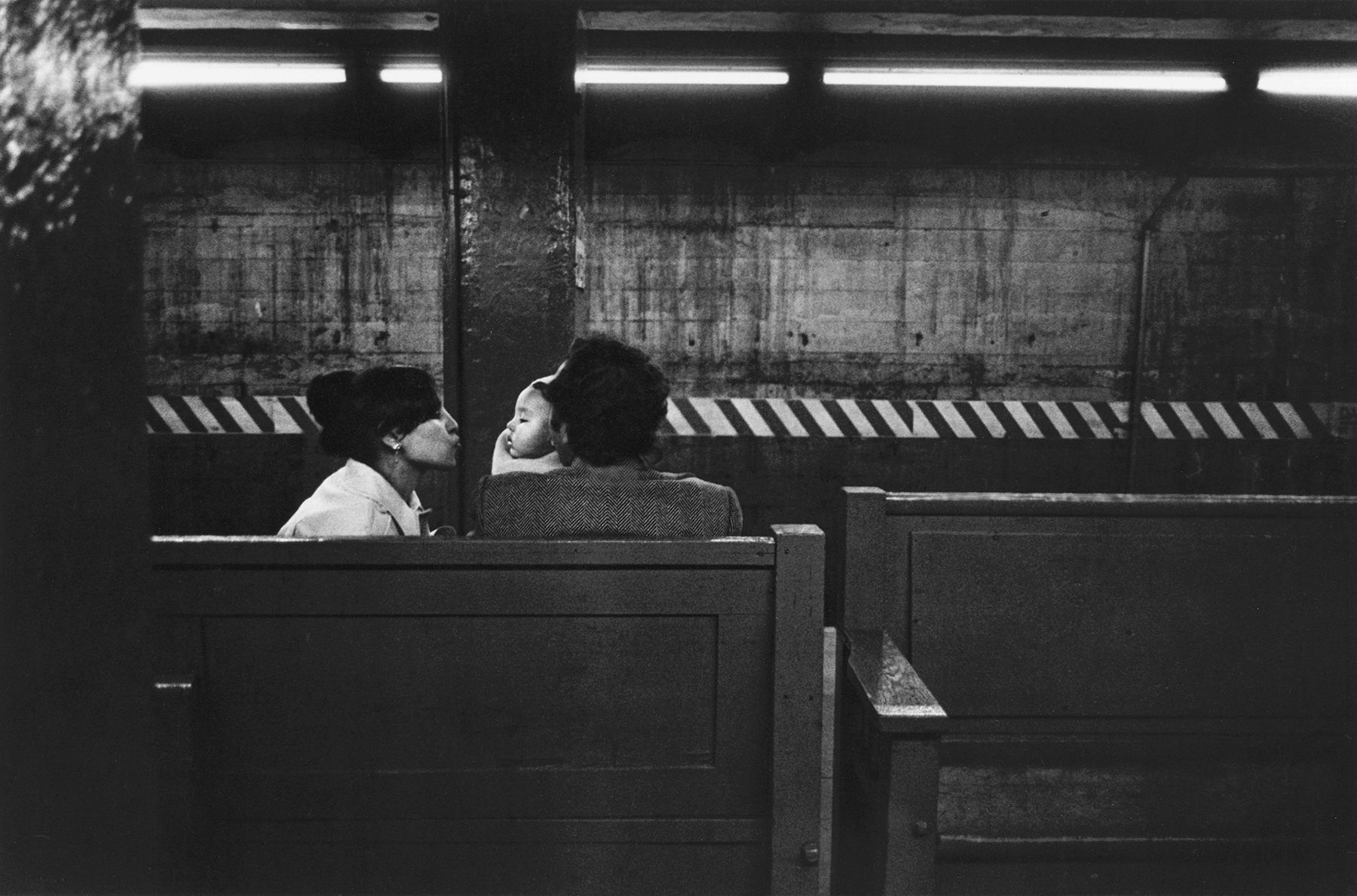Two people sitting on a bench in a dimly lit, industrial-looking setting with striped patterned walls and overhead lights.