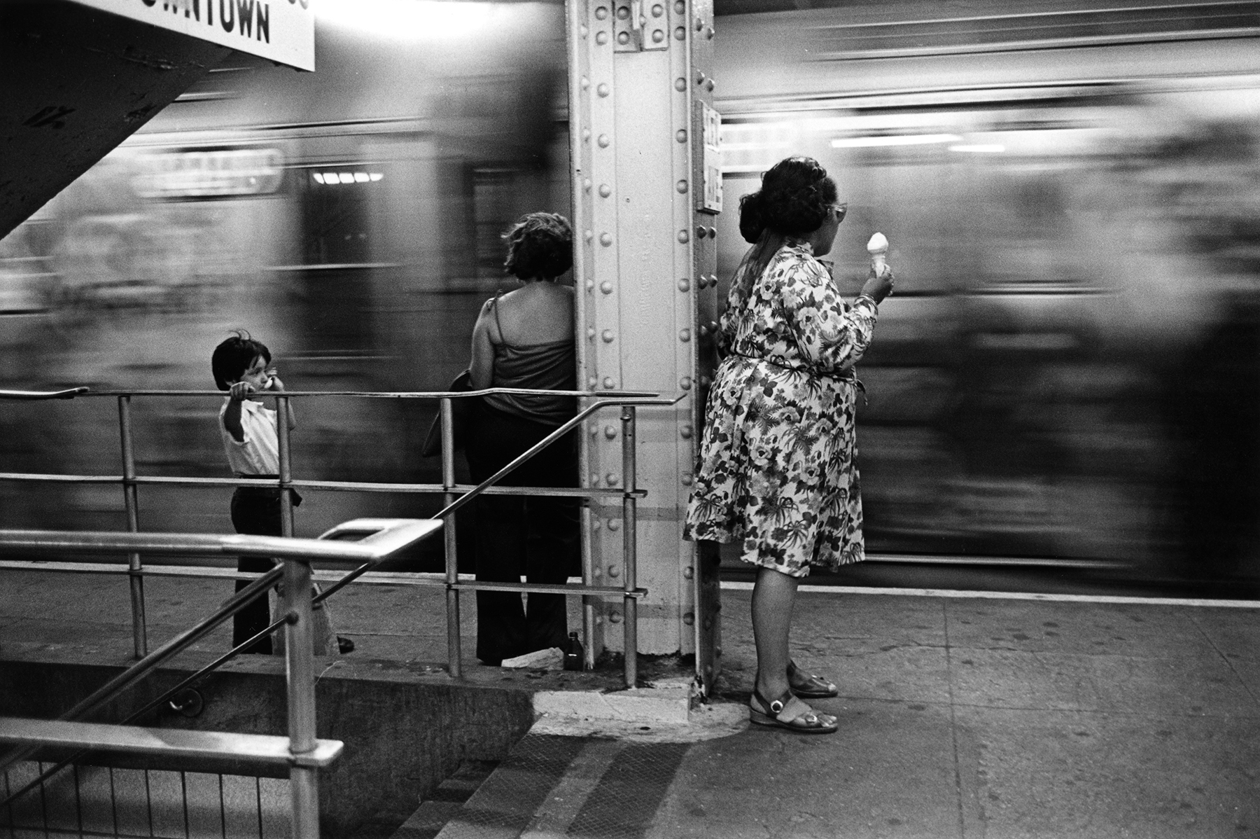 Three people standing on platform, one in a floral dress, with train speeding in background.