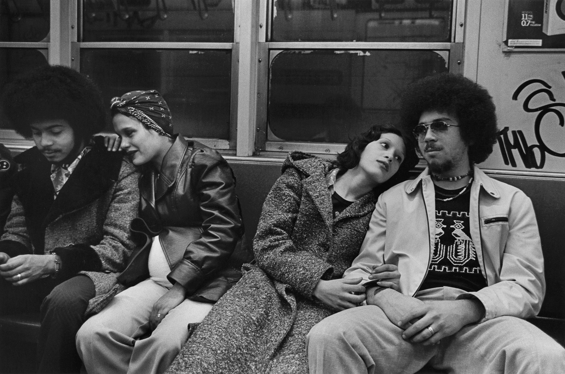 Black and white image showing four people sitting on a bench in a subway carriage. Two men and two women are visible, with the women sitting closely together in the centre of the frame.