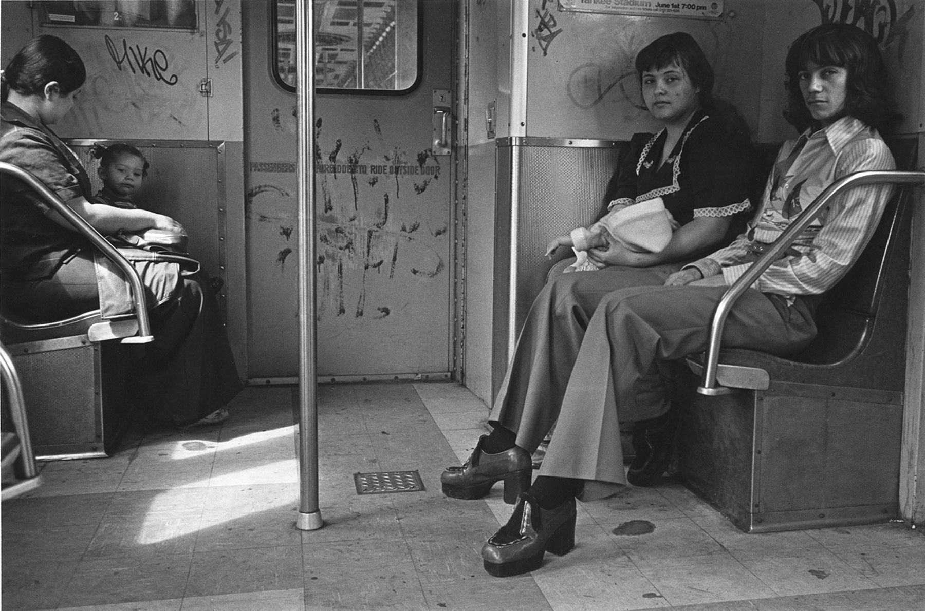Grainy monochrome image of a crowded underground train carriage, with several people sitting and standing. Two women on the right wearing long skirts and boots.