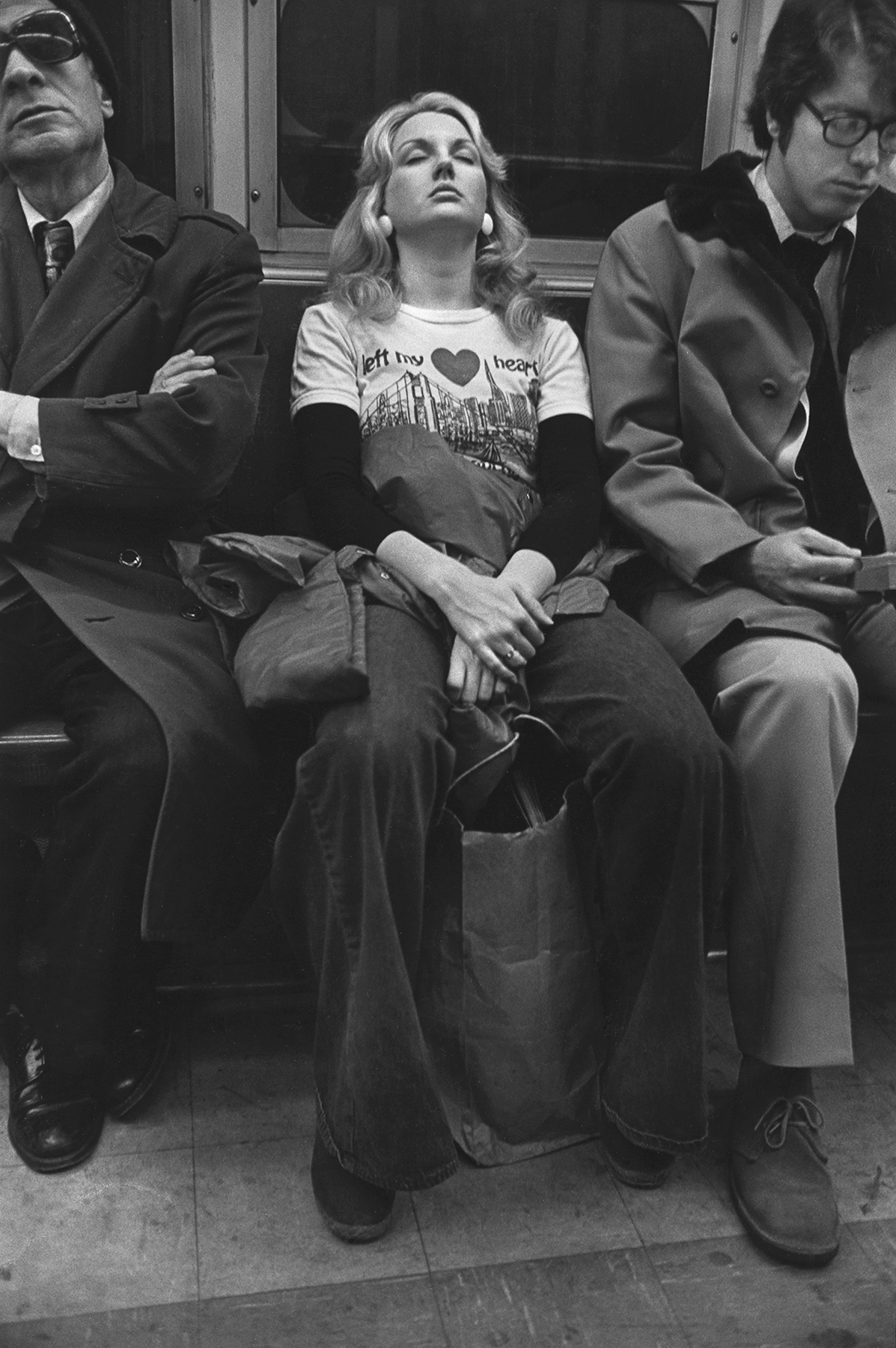 Black and white image of three people, including a woman in casual clothing, sitting on a bench.