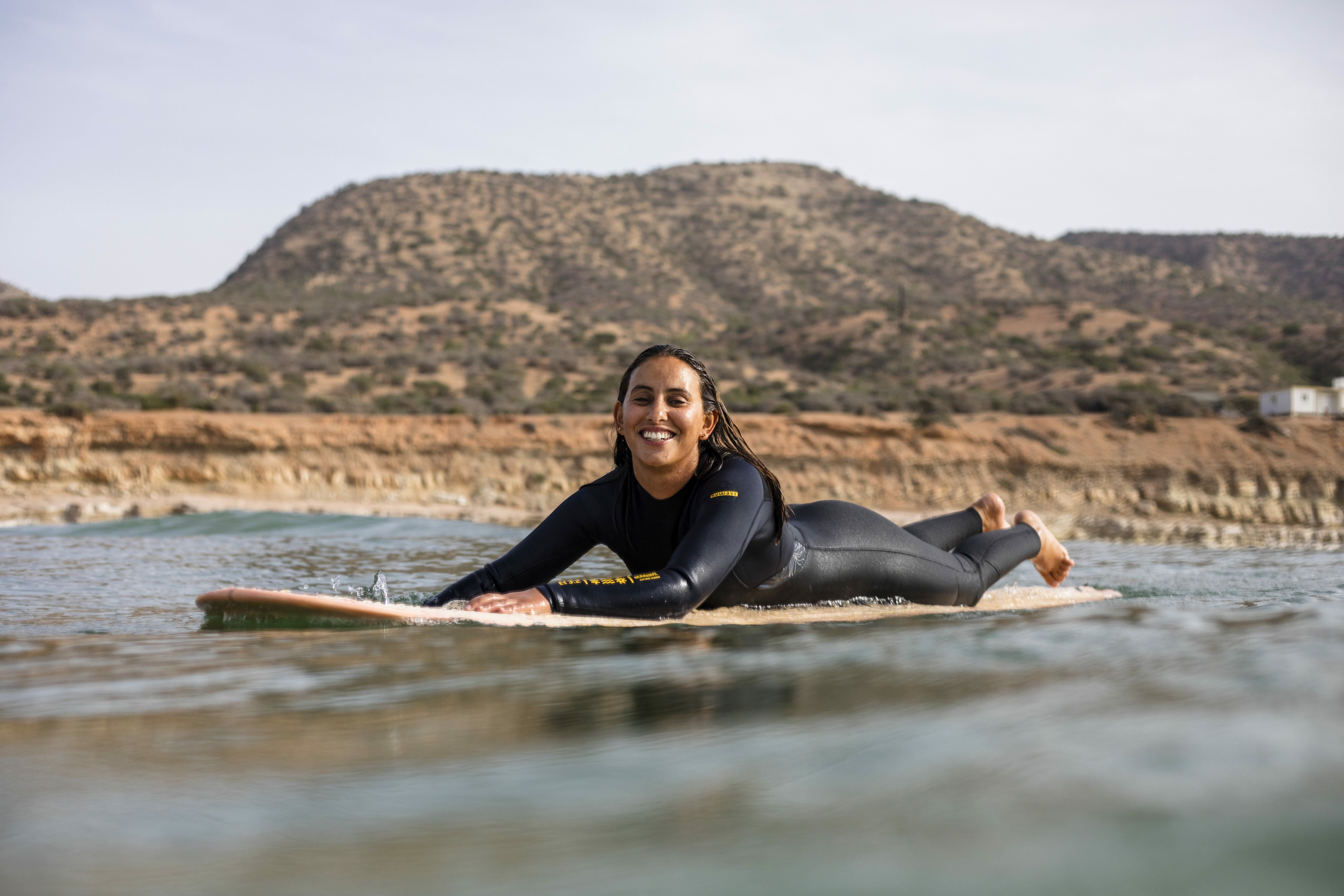 Smiling person in black wetsuit riding surfboard on calm ocean with rocky hills in background.