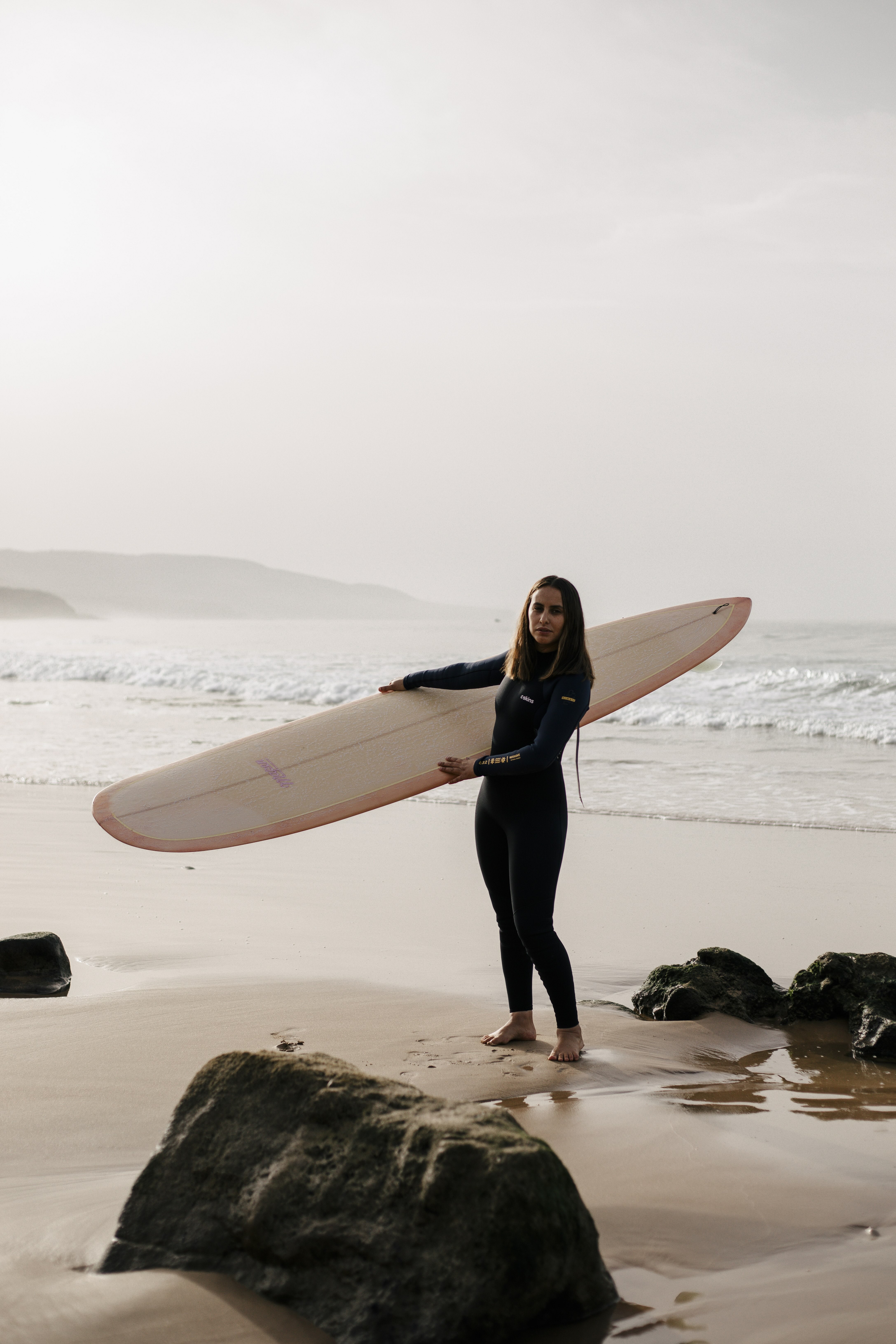 A woman in a black wetsuit holding a surfboard on a beach with rocky outcroppings, ocean and cloudy sky in the background.